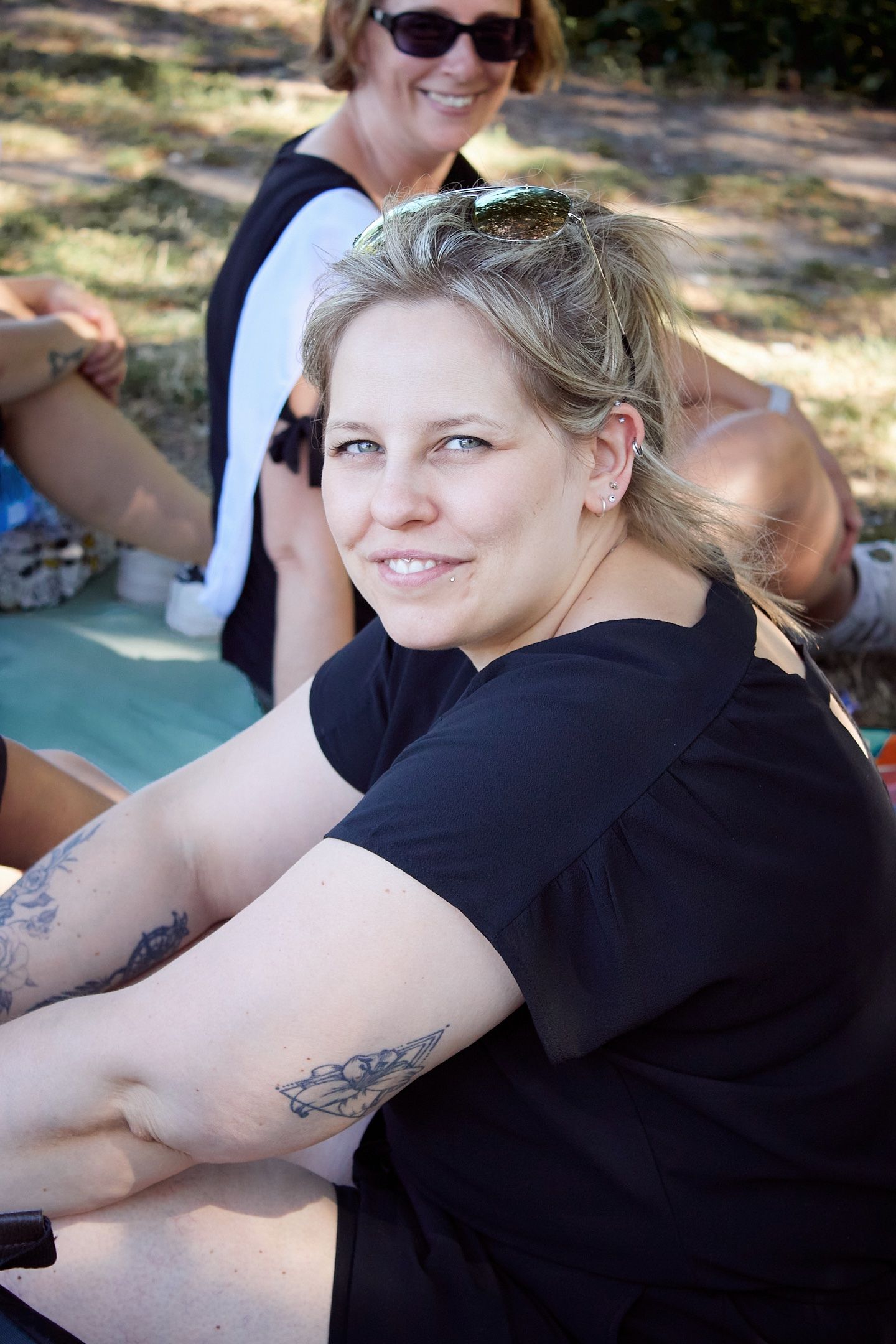 Smiling blonde woman with tattoos wearing black shirt sits outdoors at a casual gathering.