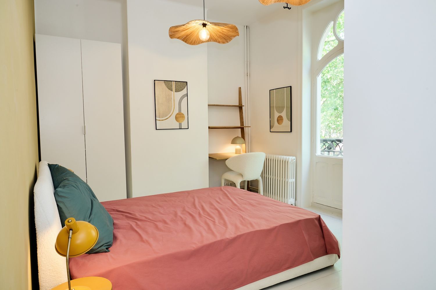 Bright bedroom with pink bedspread, white wardrobe, wooden desk, and large window with natural light.