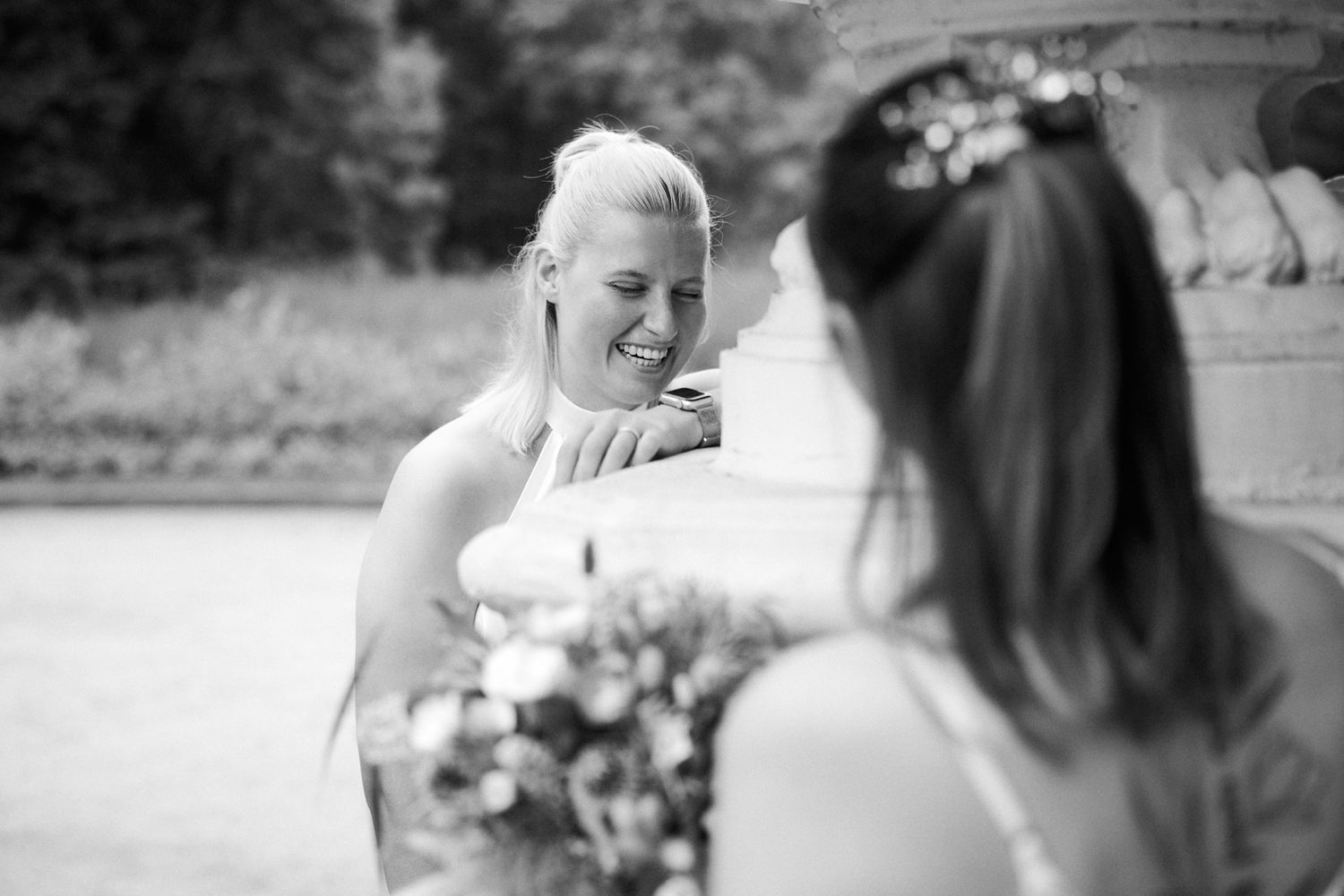 Two brides share a joyful moment outdoors, one smiling warmly while holding a bouquet, in a black and white wedding photo.