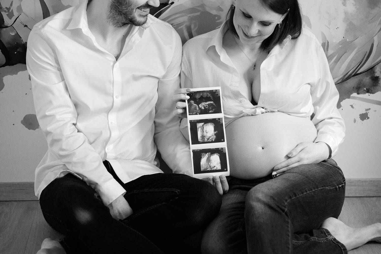 Black and white photo of pregnant couple holding ultrasound scans, woman showing baby bump, sitting together.