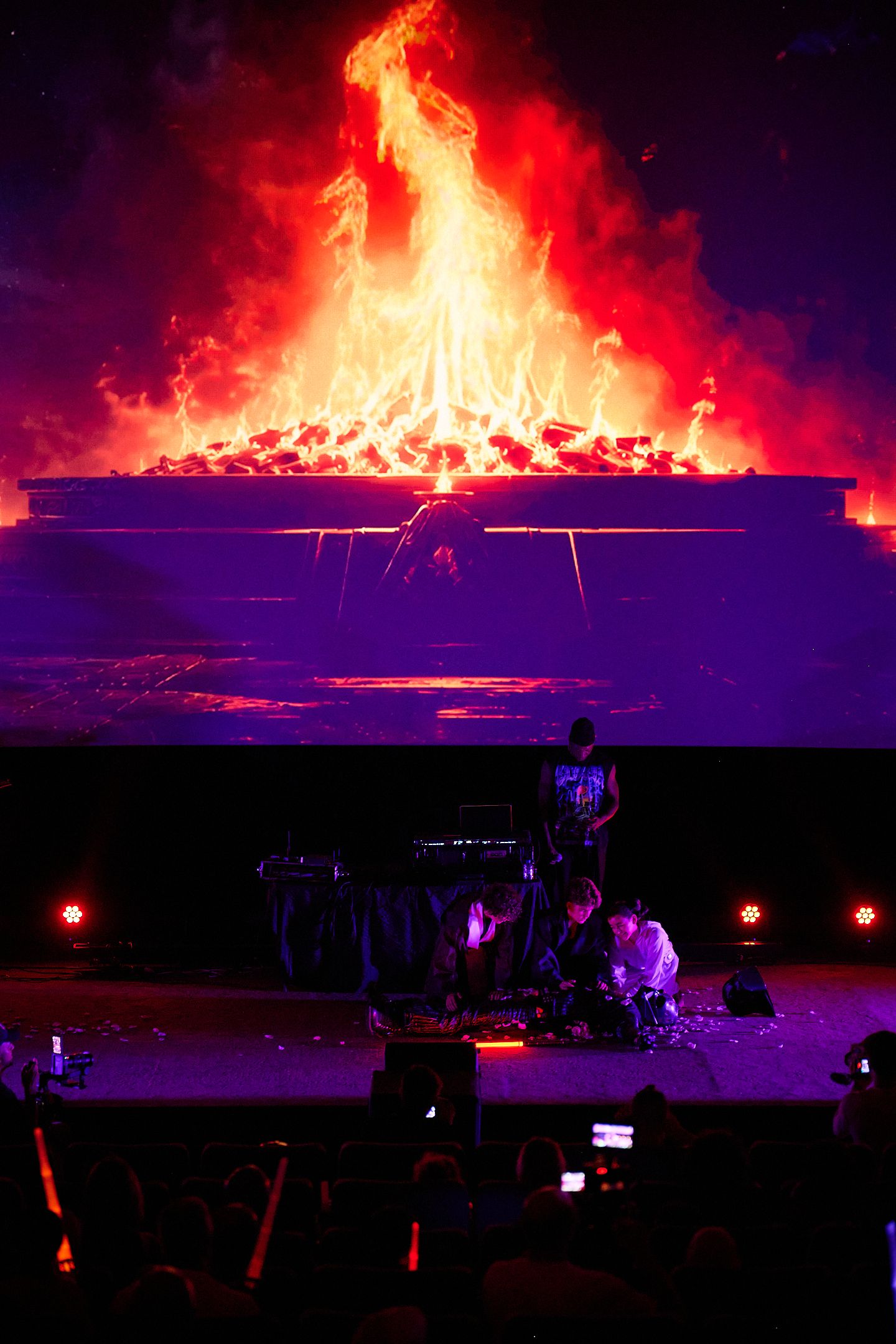 Concert stage with dramatic fire backdrop display, performers on purple-lit stage with audience silhouettes in foreground.
