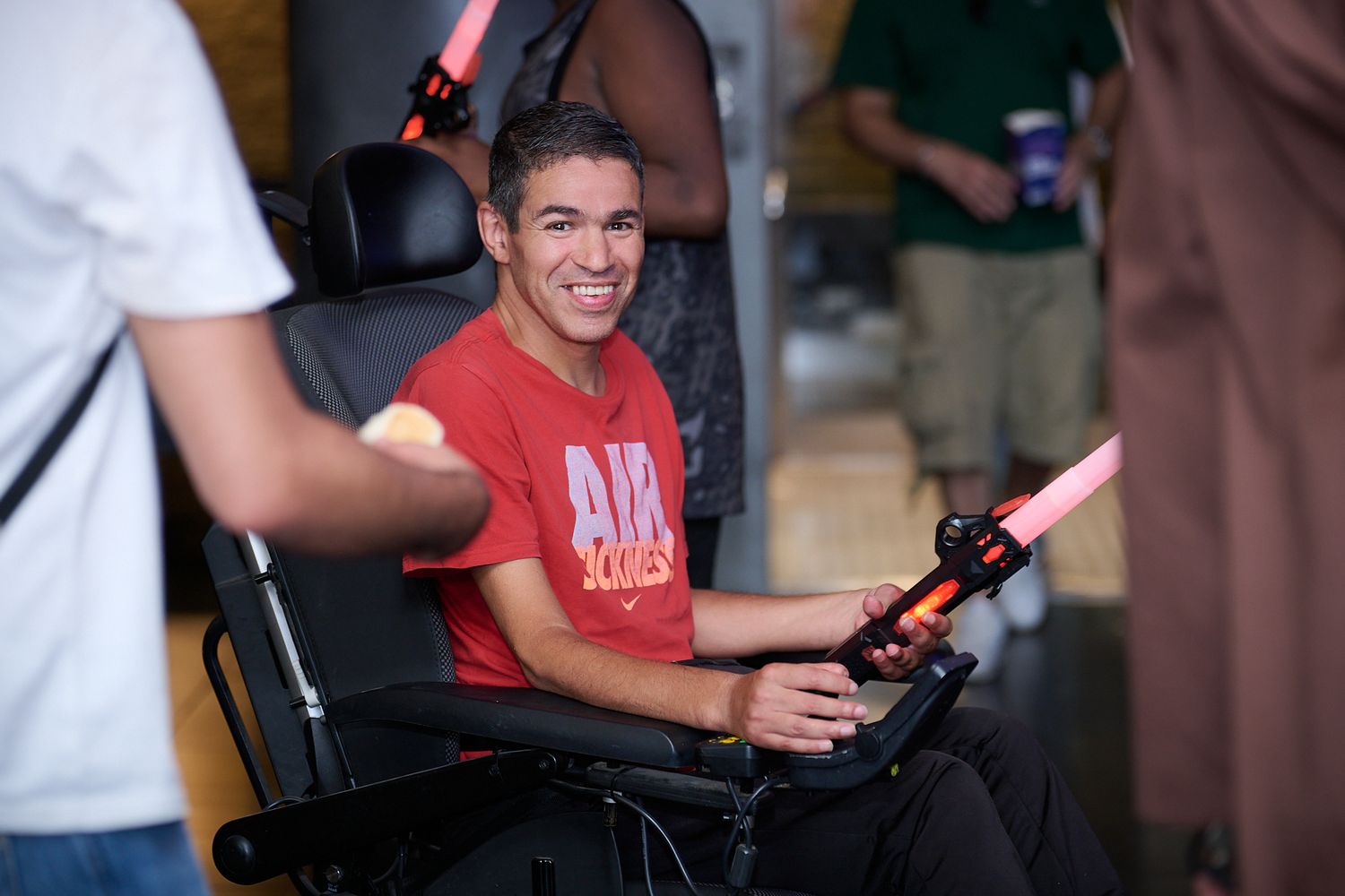 Man in red Air shirt smiling in wheelchair, holding pink laser tag gun at indoor gaming event.