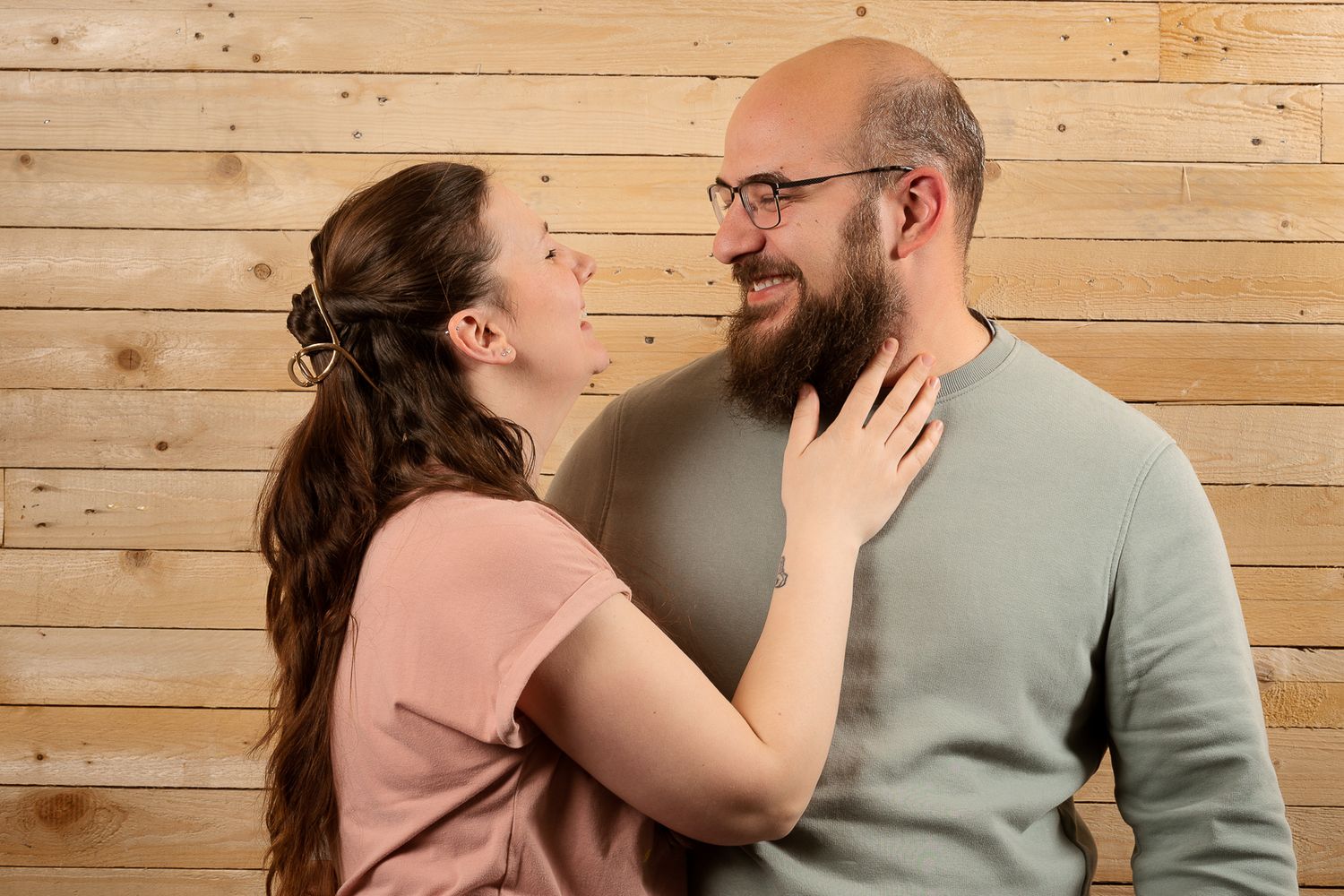 Affectionate couple smiling at each other, woman touching mans beard against wooden wall background.