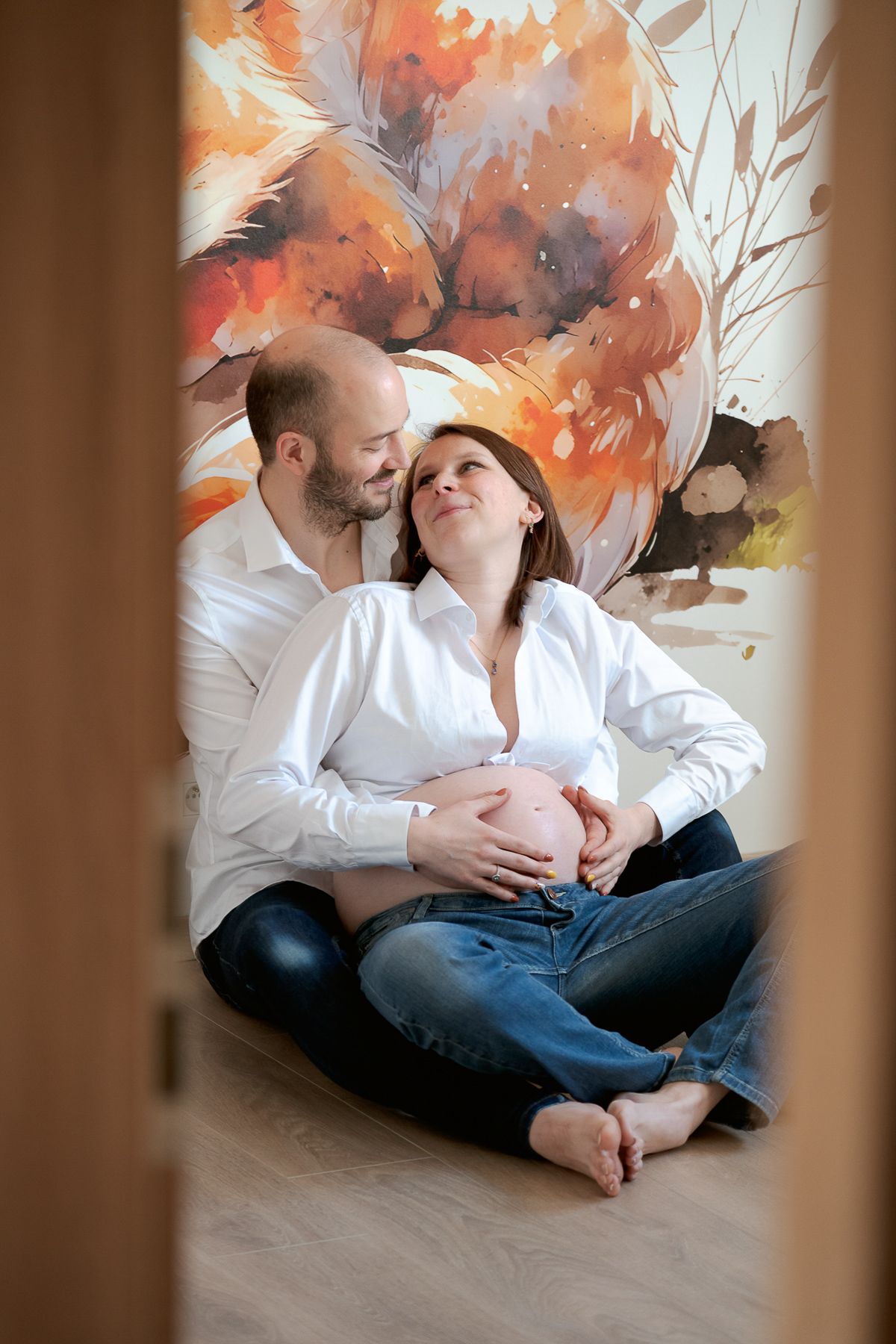 Couple sitting on floor, pregnant woman cradling baby bump, partner embracing her, colorful floral mural backdrop.