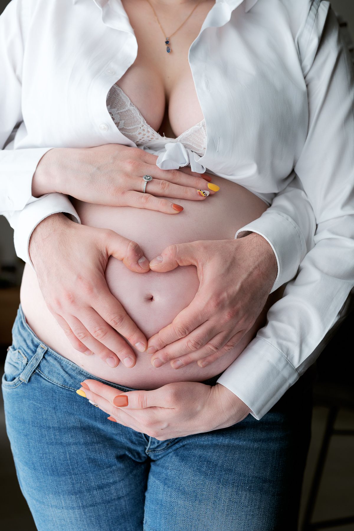 Couple forming heart shape with hands on pregnant womans bare belly, white shirt open.