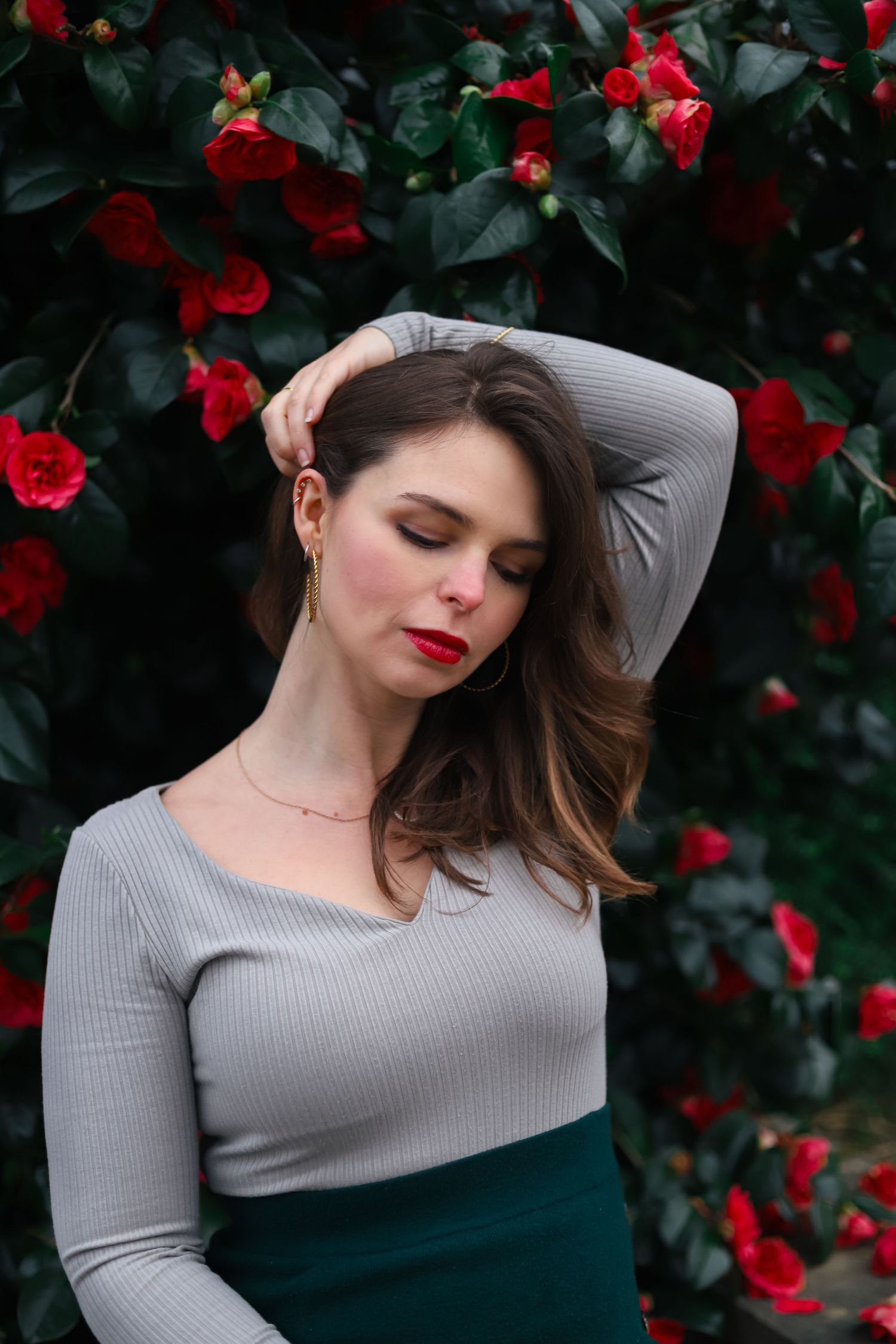 Elegant woman in grey top and green skirt with eyes closed beside blooming red camellia bushes.