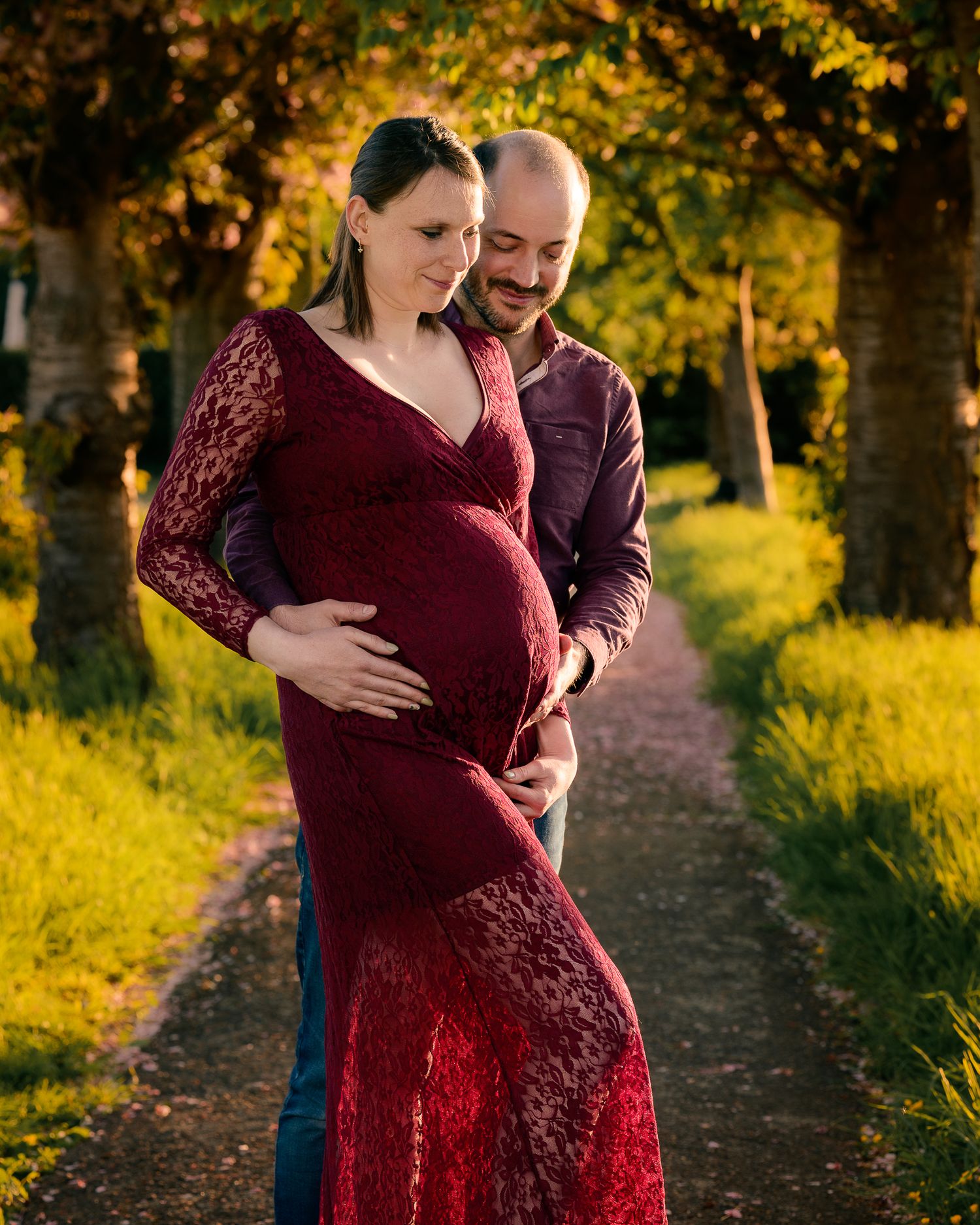 Couple embracing on a tree-lined path at golden hour, woman wearing a red lace maternity gown.