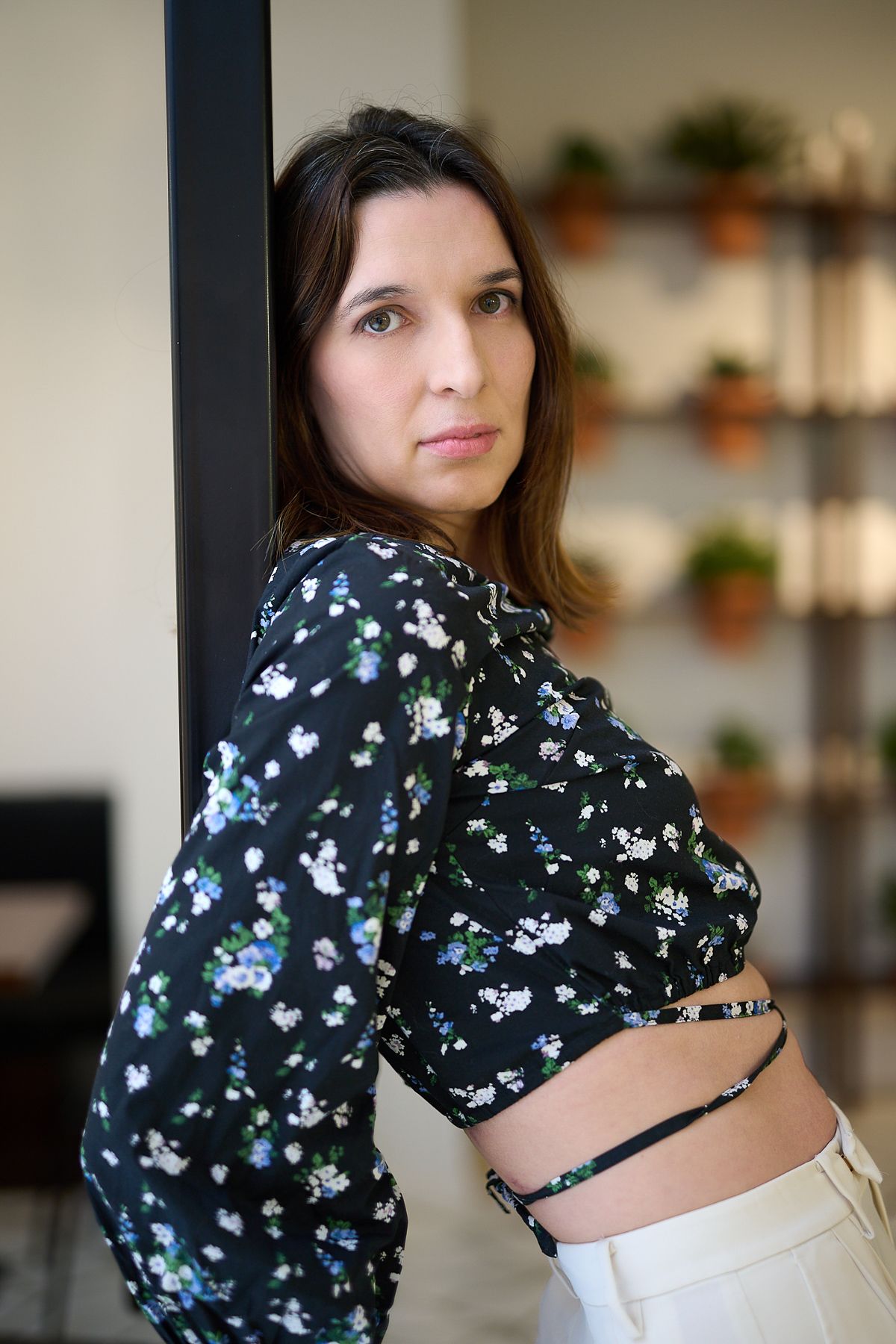 Woman in black floral crop top leaning against a dark pillar, with shelves of potted plants in the background.