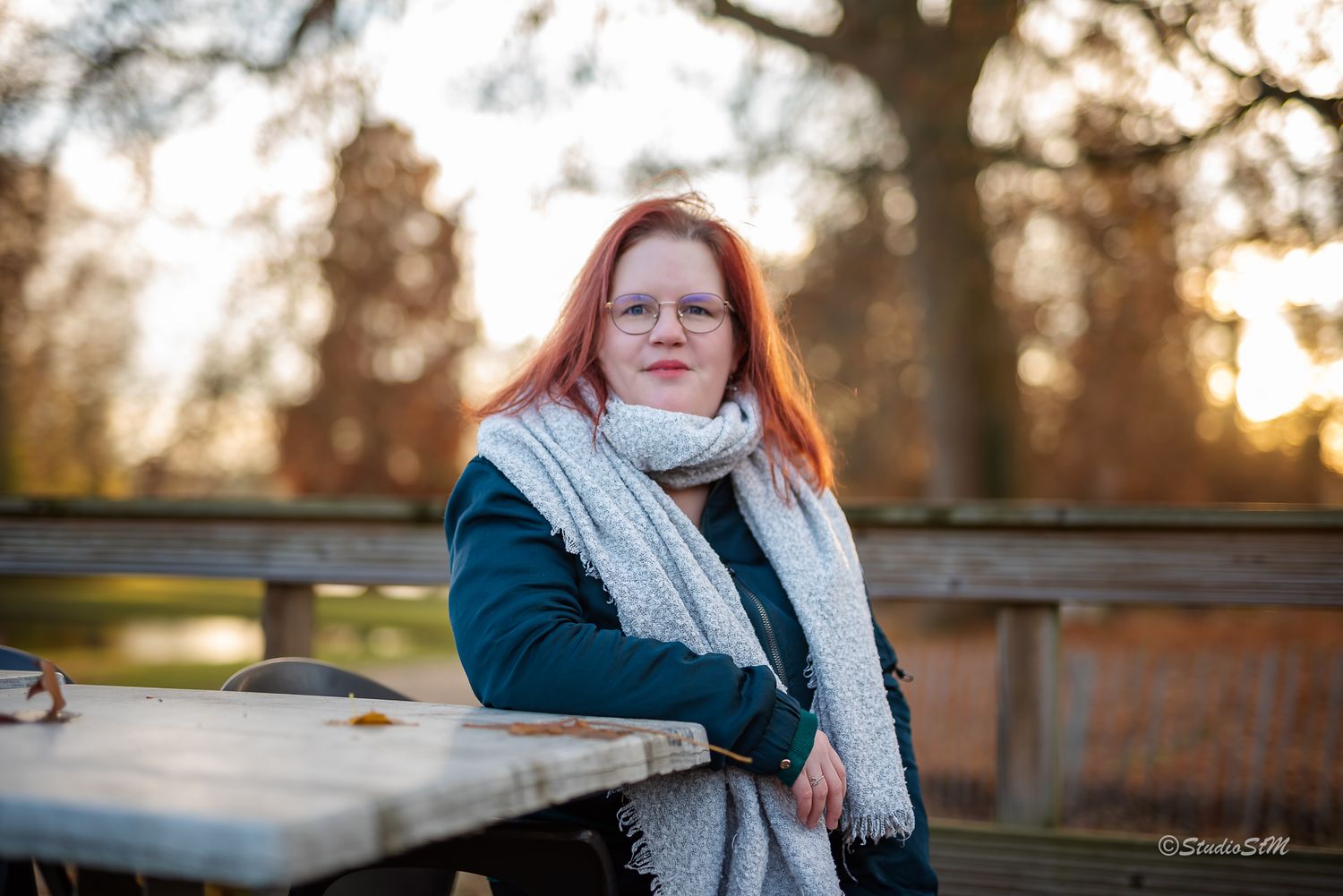 Red-haired woman in glasses and white scarf leans on a wooden table in a sunny park.