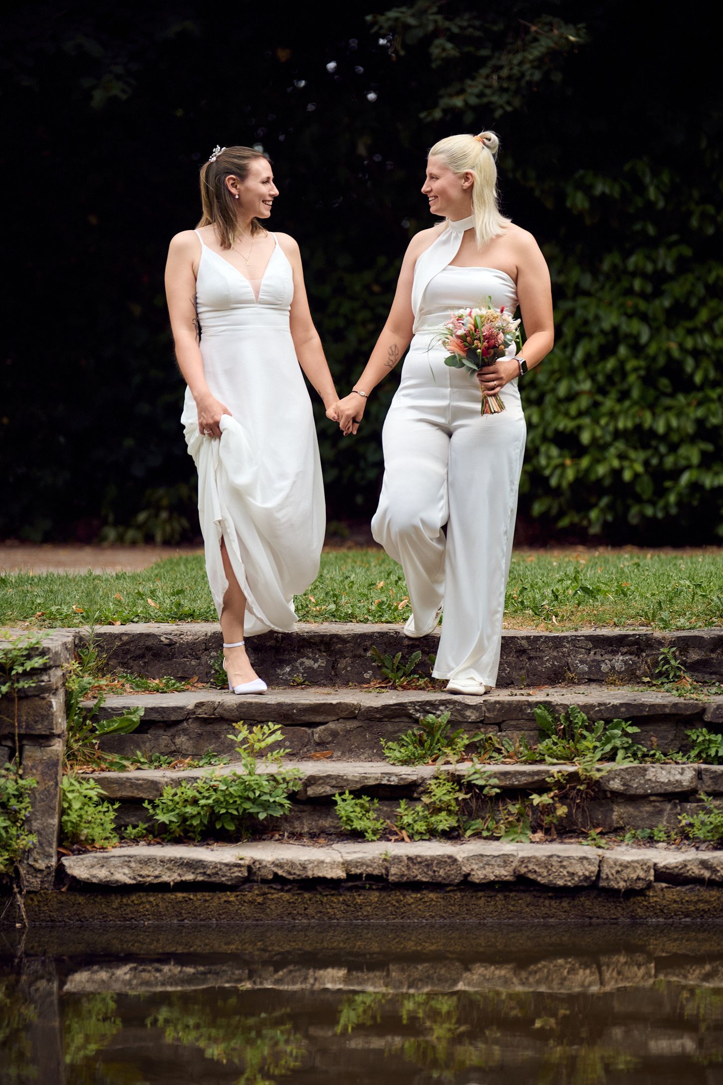 Two brides in white holding hands and bouquet on stone steps outdoors.