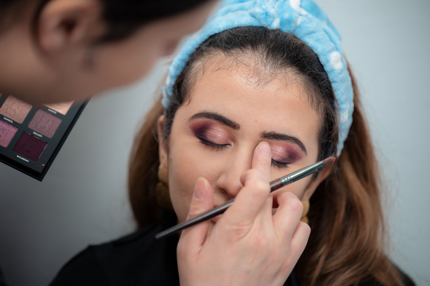 Makeup artist applying purple eyeshadow to a young woman wearing a blue headband.