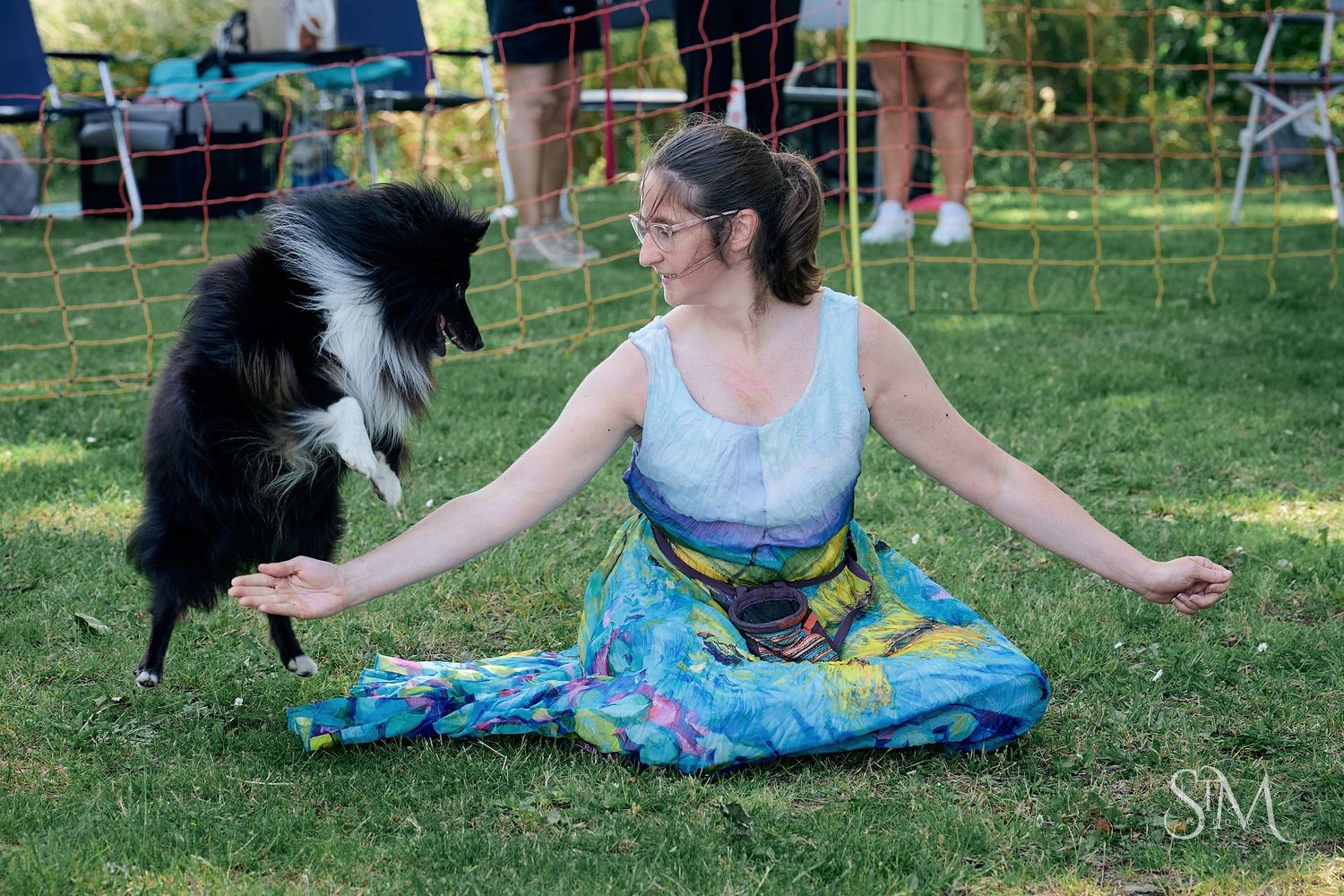 Woman in colorful dress meditates on grass while a border collie dog interacts with her outdoors.