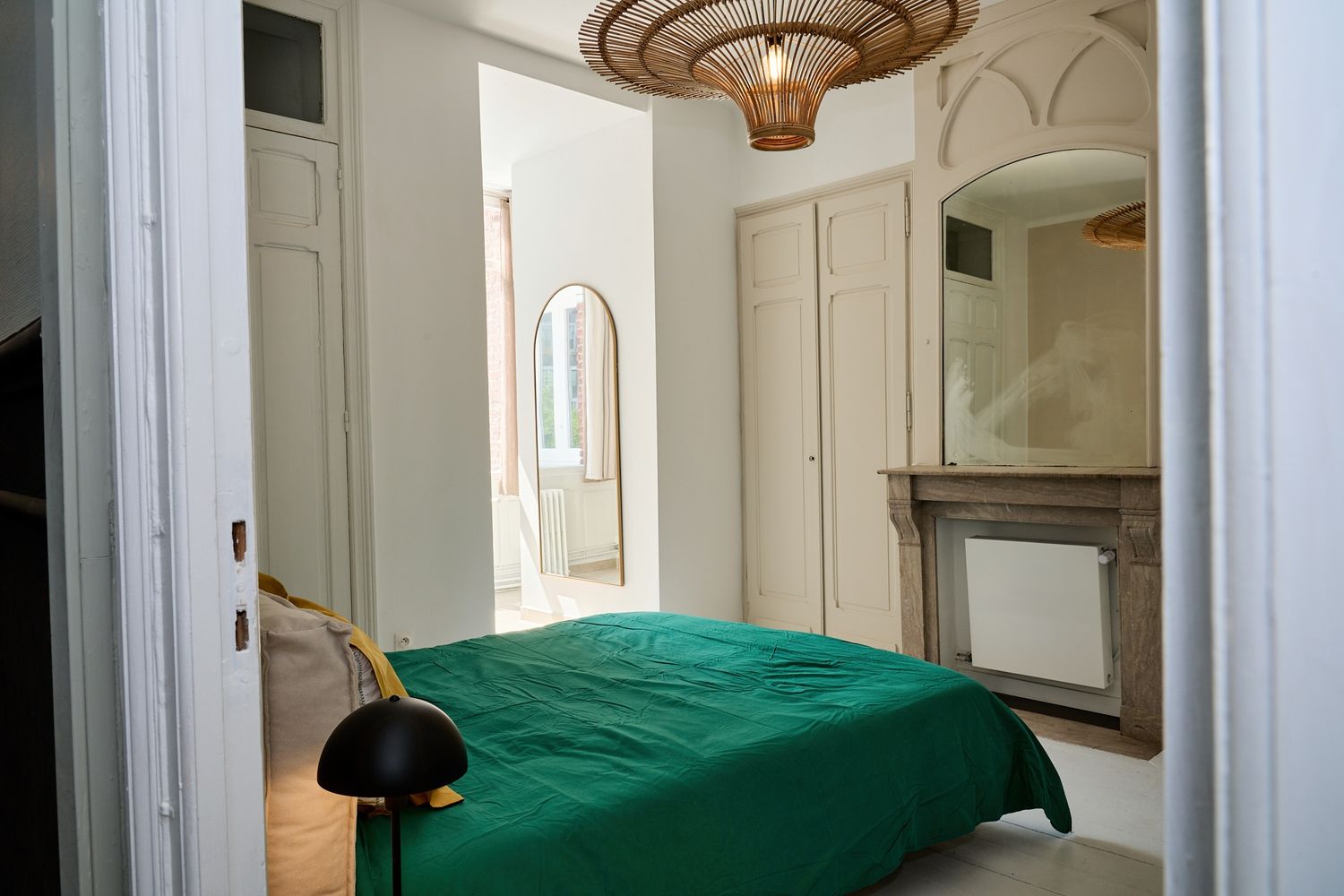 Elegant bedroom with green bedspread, fireplace, arched doorway, and rattan pendant light in a bright white room.