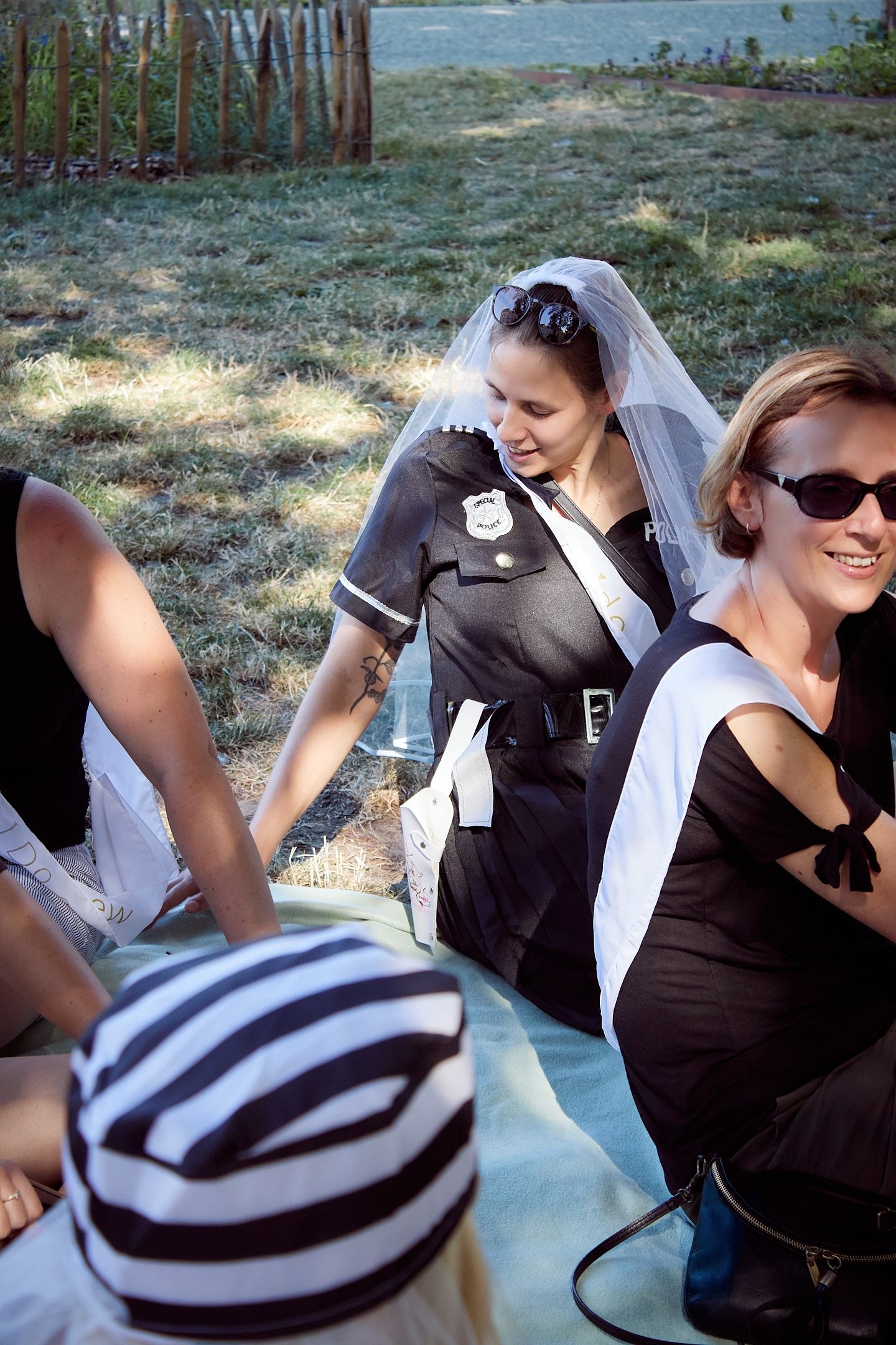 Woman in police costume with bridal veil at outdoor bachelorette party, sitting with friends in black dresses.