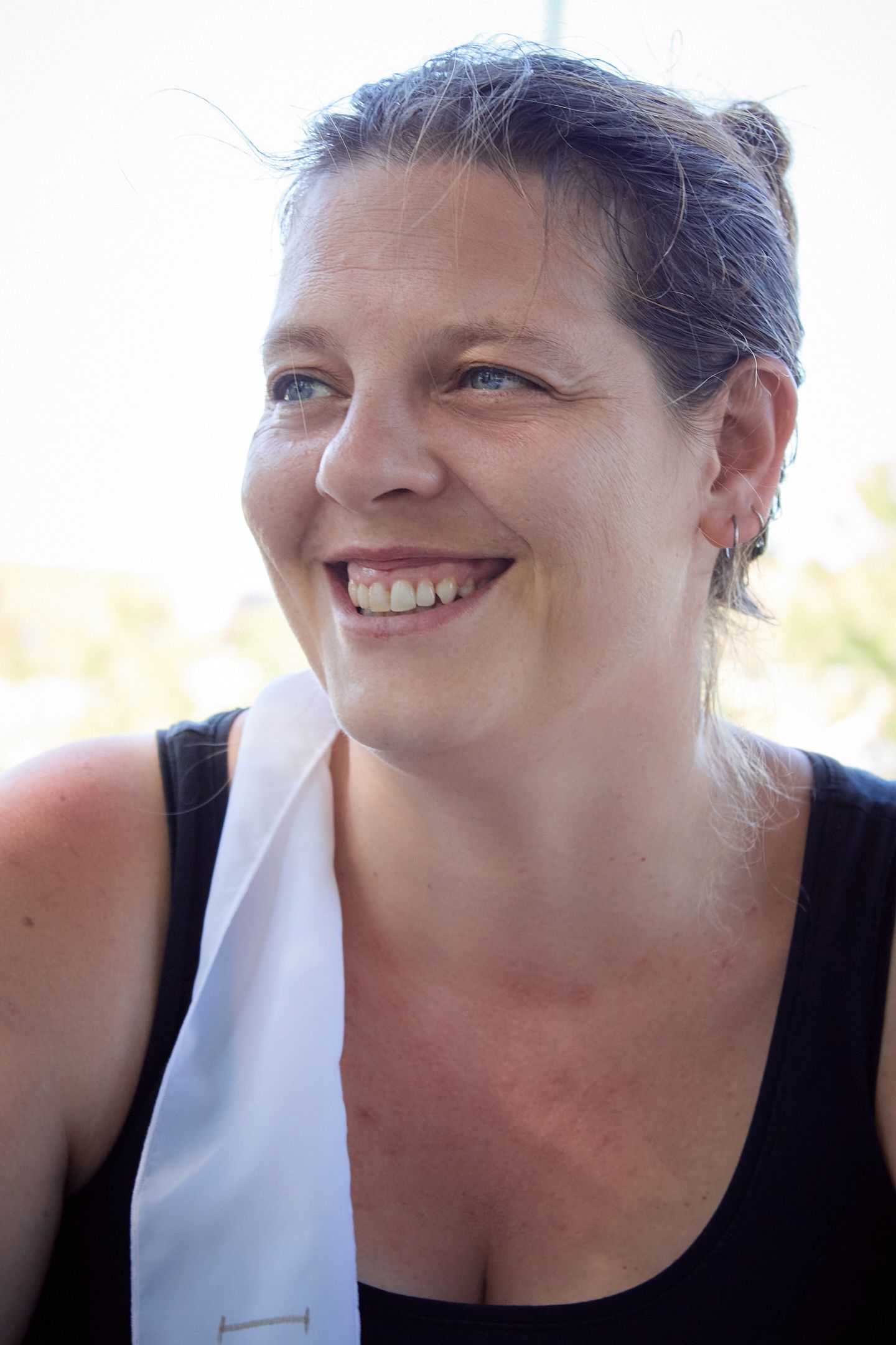 Smiling middle-aged woman with blue eyes wearing a black tank top and white scarf outdoors.