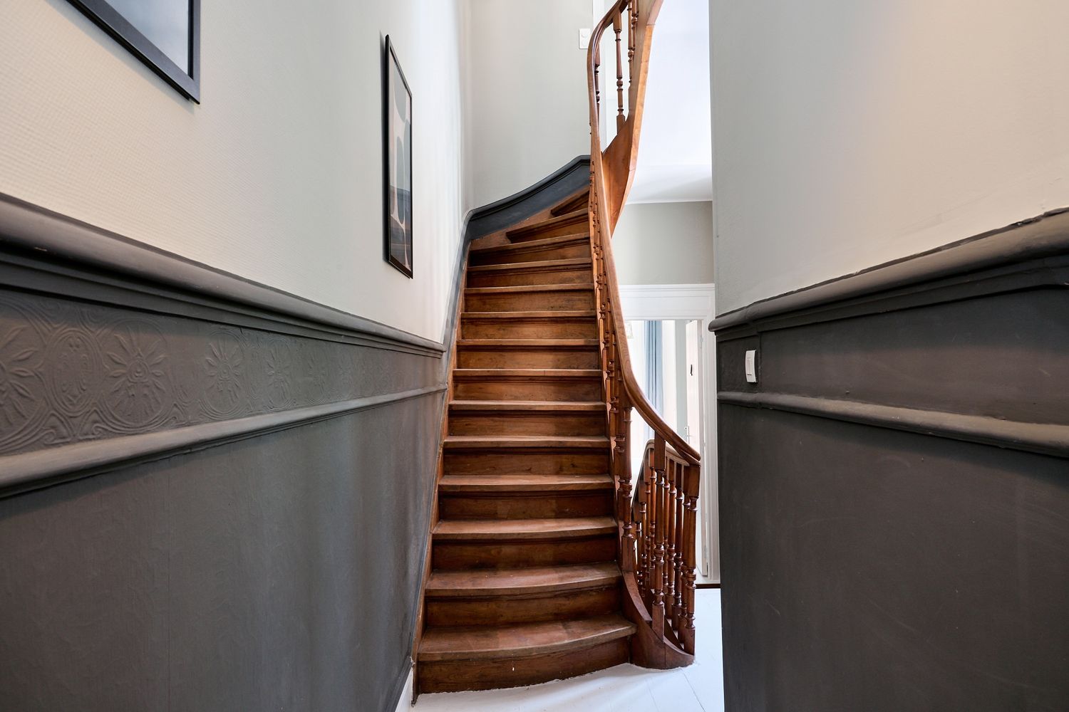 Elegant wooden staircase with curved banister in a hallway featuring gray wainscoting and white walls.