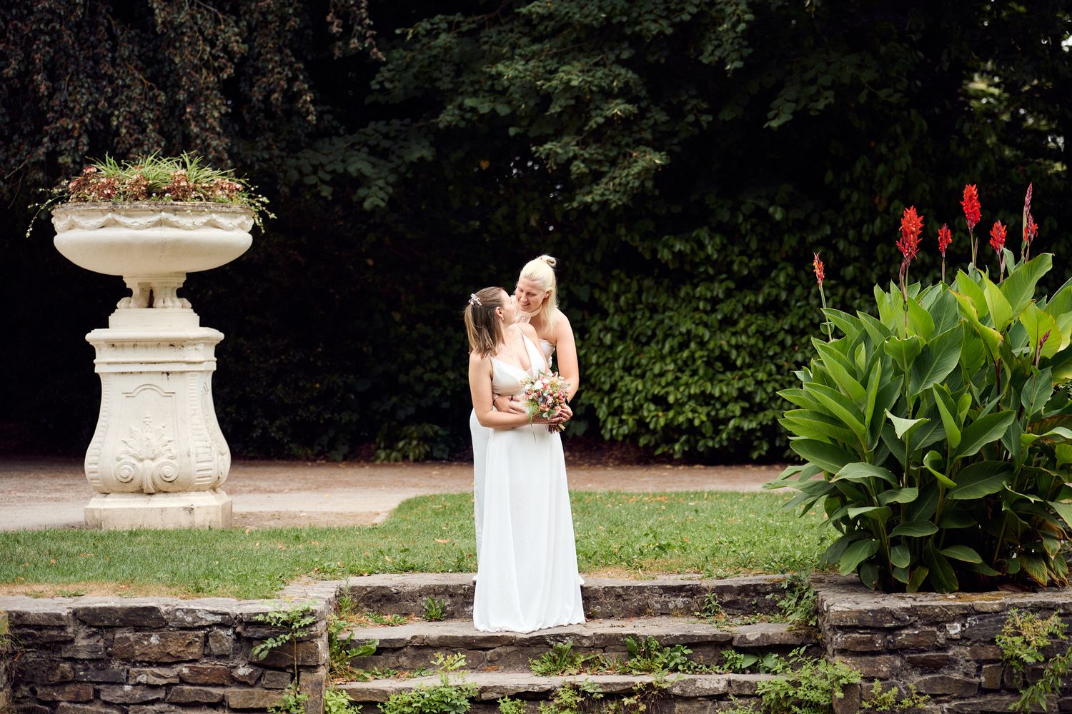 Two brides share a romantic kiss in a lush garden setting with stone fountain and red flowers.