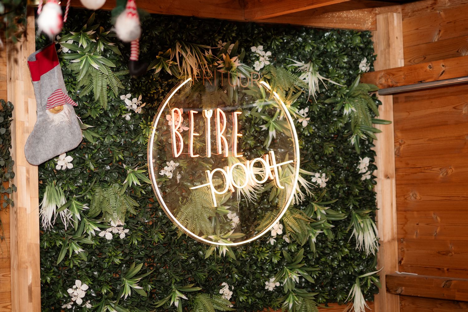 Neon sign reading BeYBE pool on a lush green foliage wall with white flowers and holiday stocking decor.
