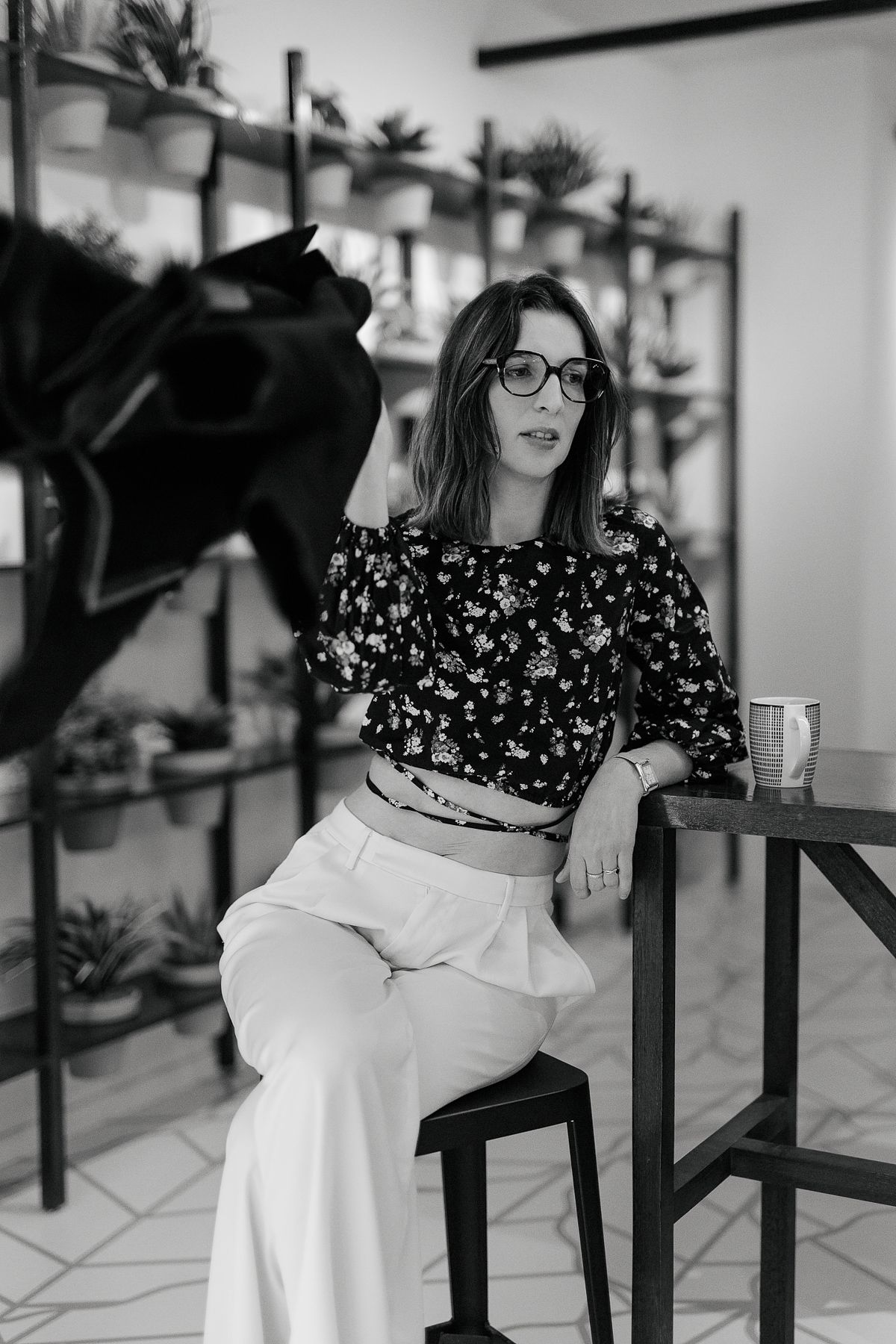 Black and white photo of a woman in floral top and white pants sitting on a stool in a plant-filled studio.