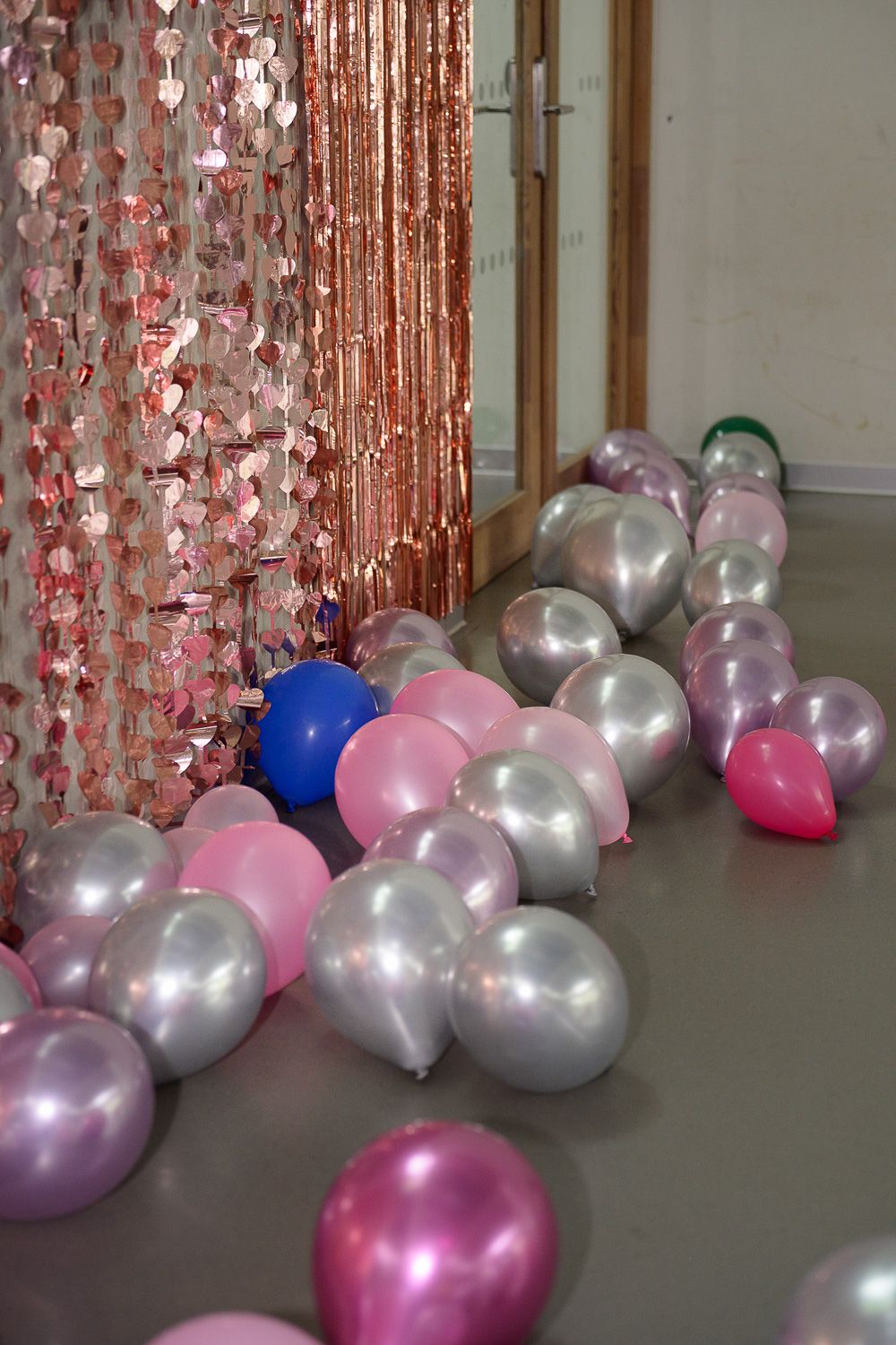 Colorful metallic balloons in pink, silver, and blue scattered on floor near rose gold tinsel curtain.
