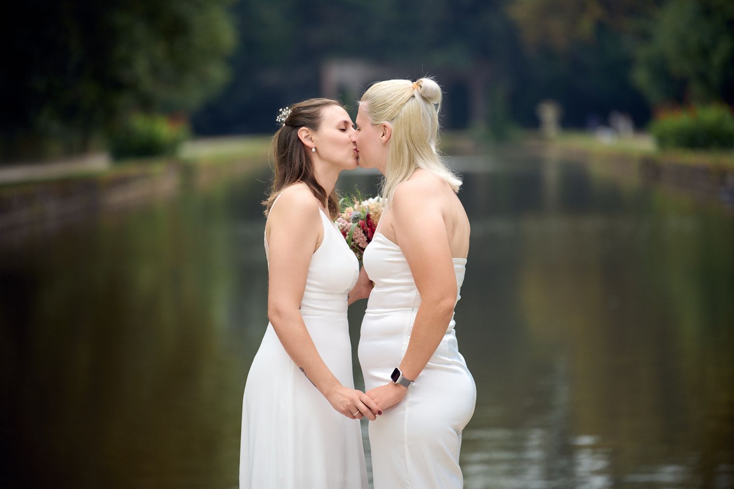 Two brides in white dresses share a kiss near a serene lake surrounded by lush greenery on their wedding day.