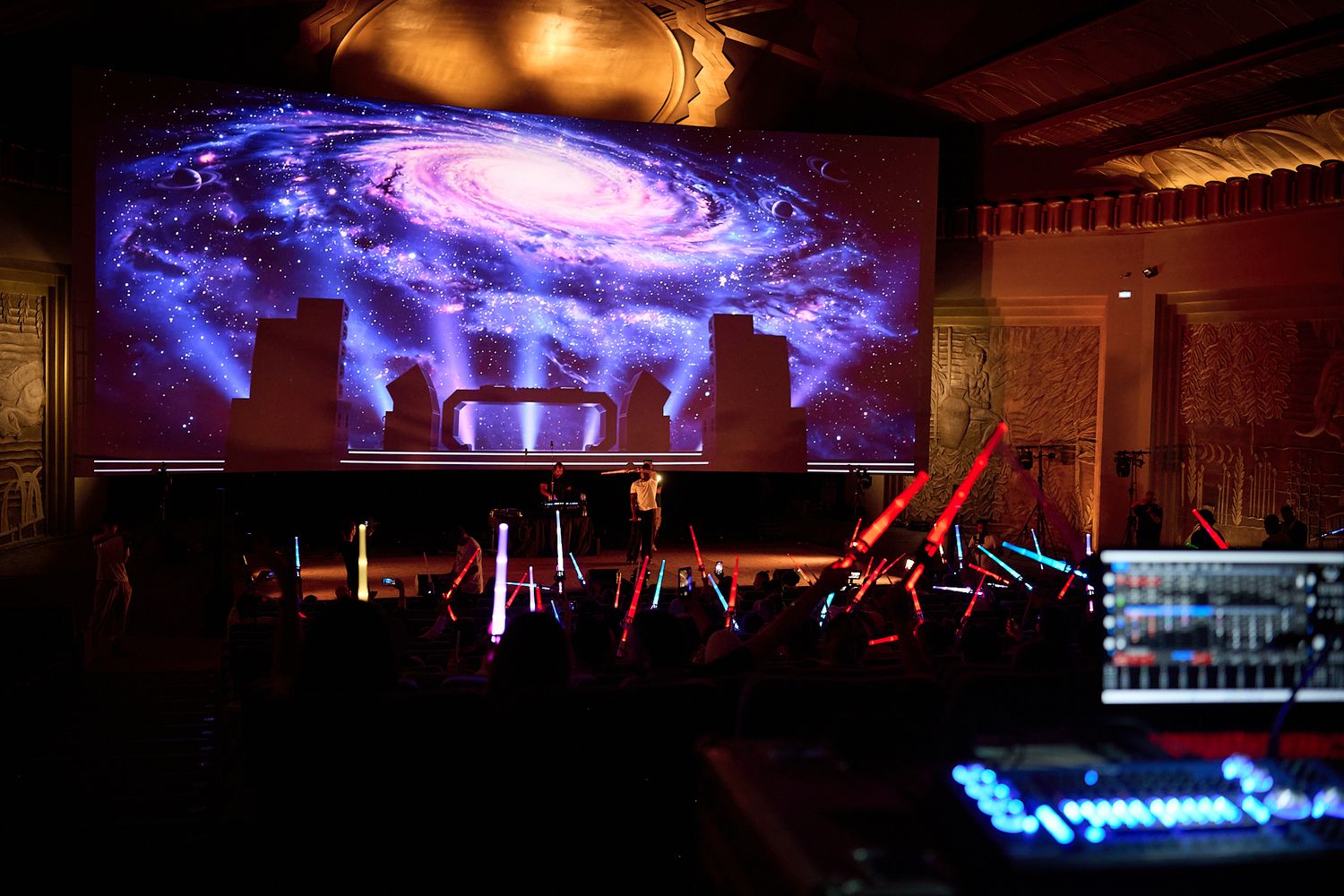 Concert venue with galaxy projection screen, silhouetted performers, glowing lightsabers, and mixing board in foreground.