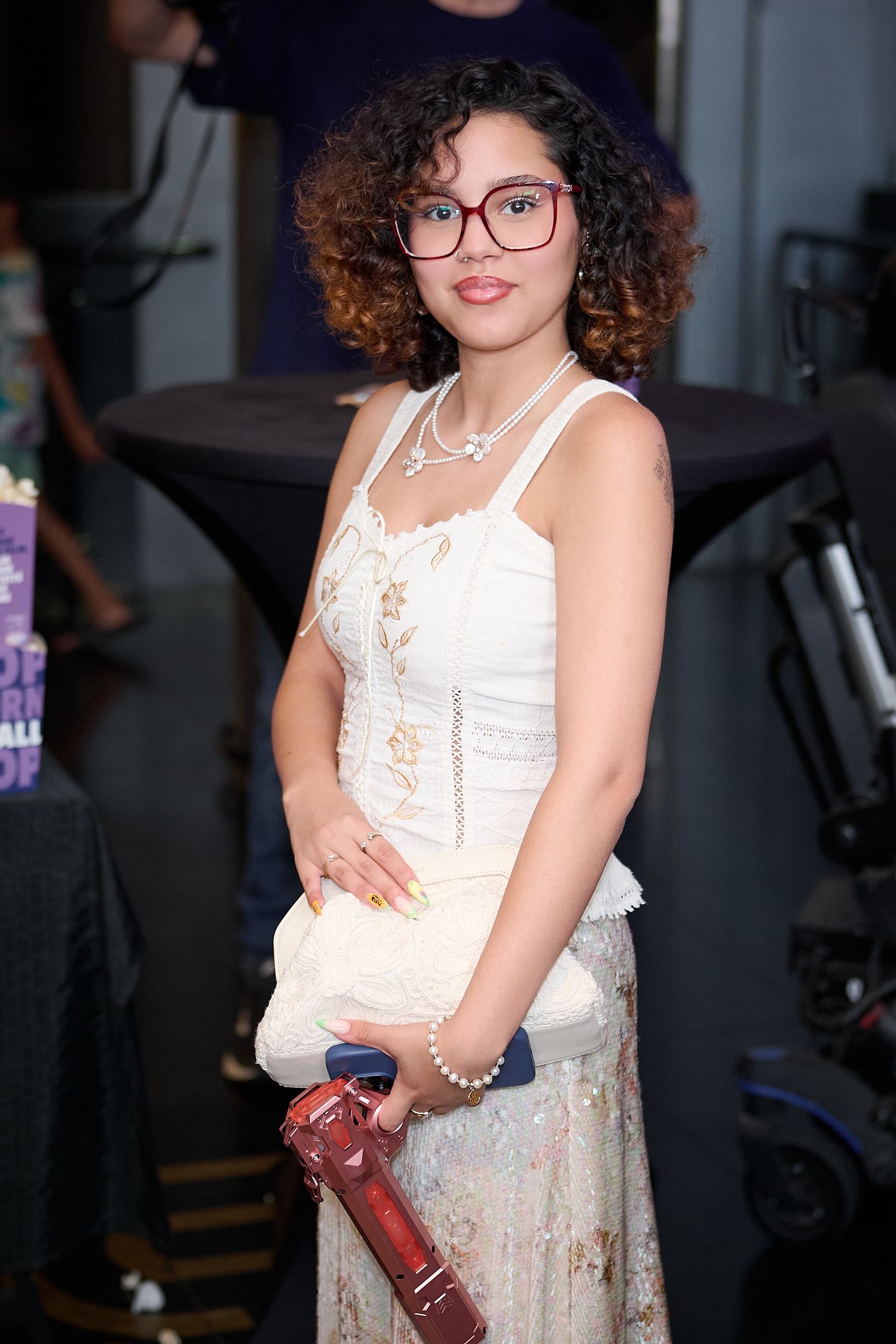 Young woman with curly hair and red glasses wearing a white lace dress, holding a handbag at an indoor event.