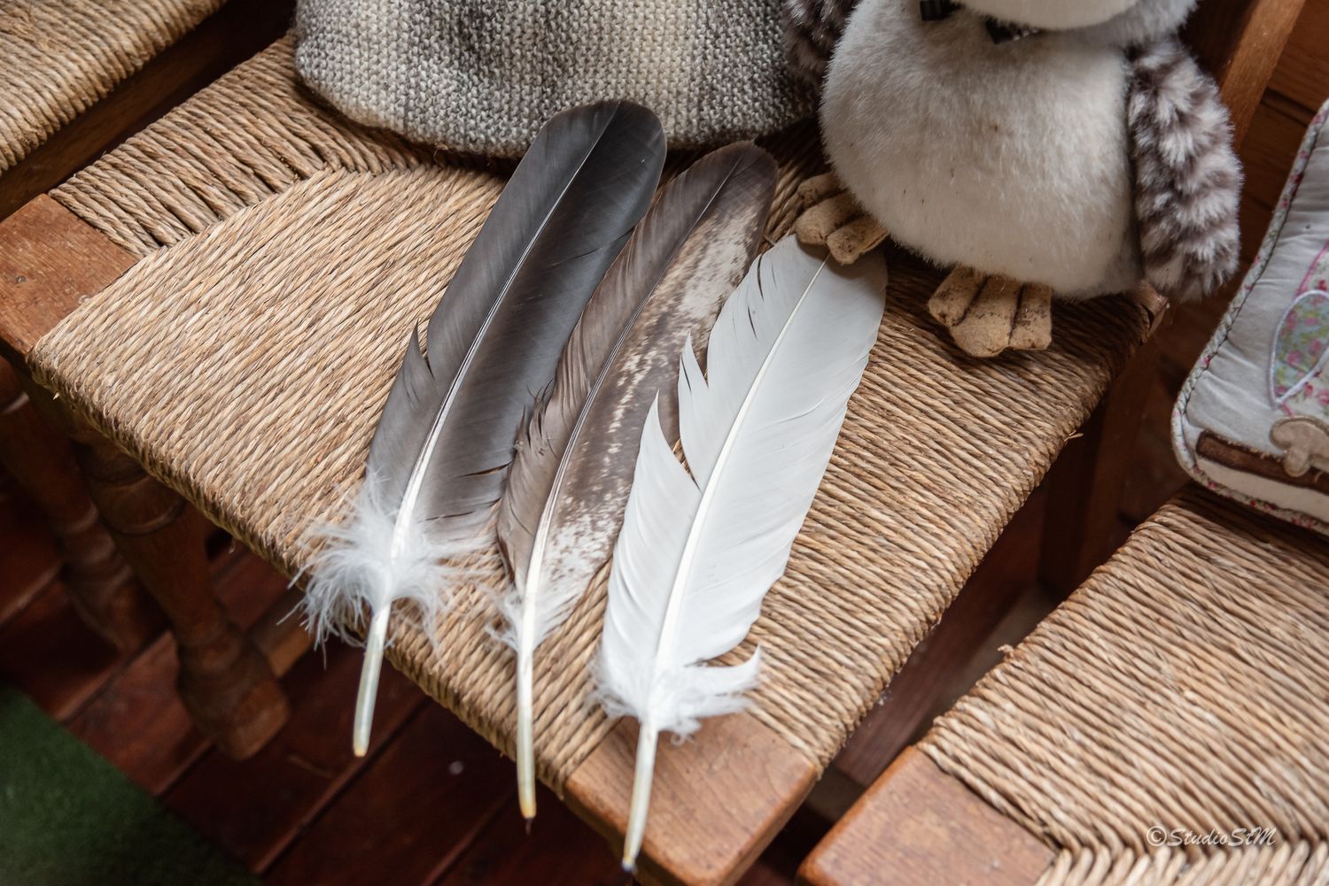 Three feathers resting on a woven wicker chair beside a small owl figurine.