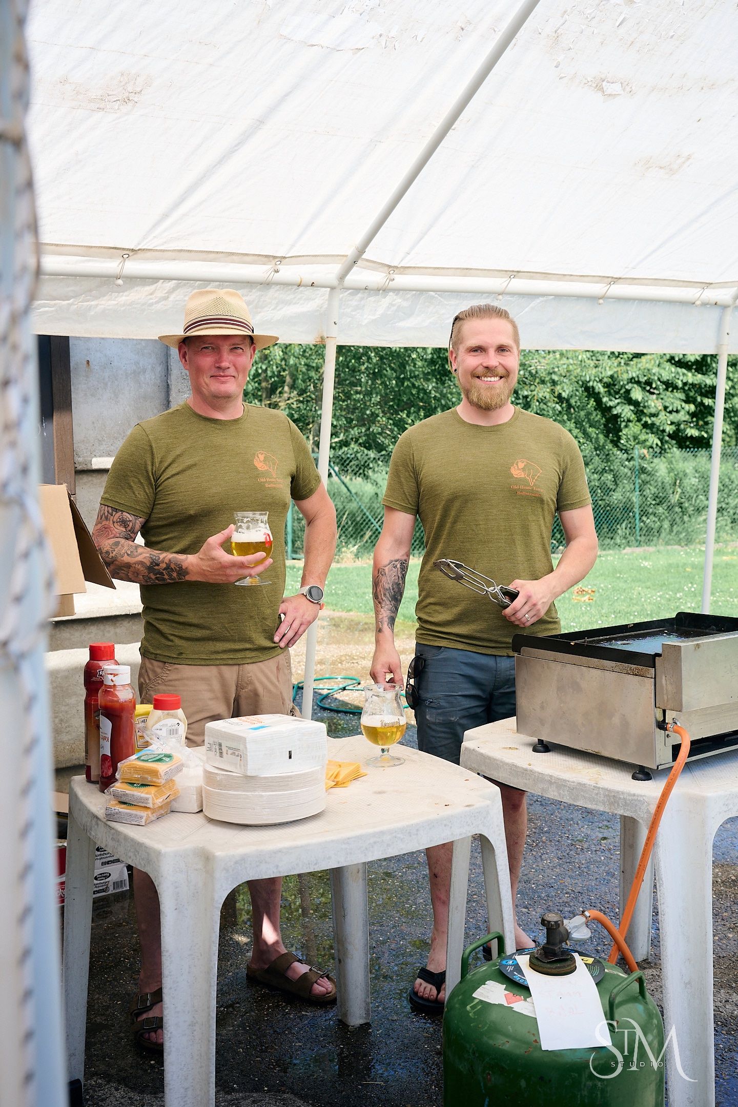 Two men in matching green t-shirts smile while grilling outdoors under a tent, one holding a beer glass.