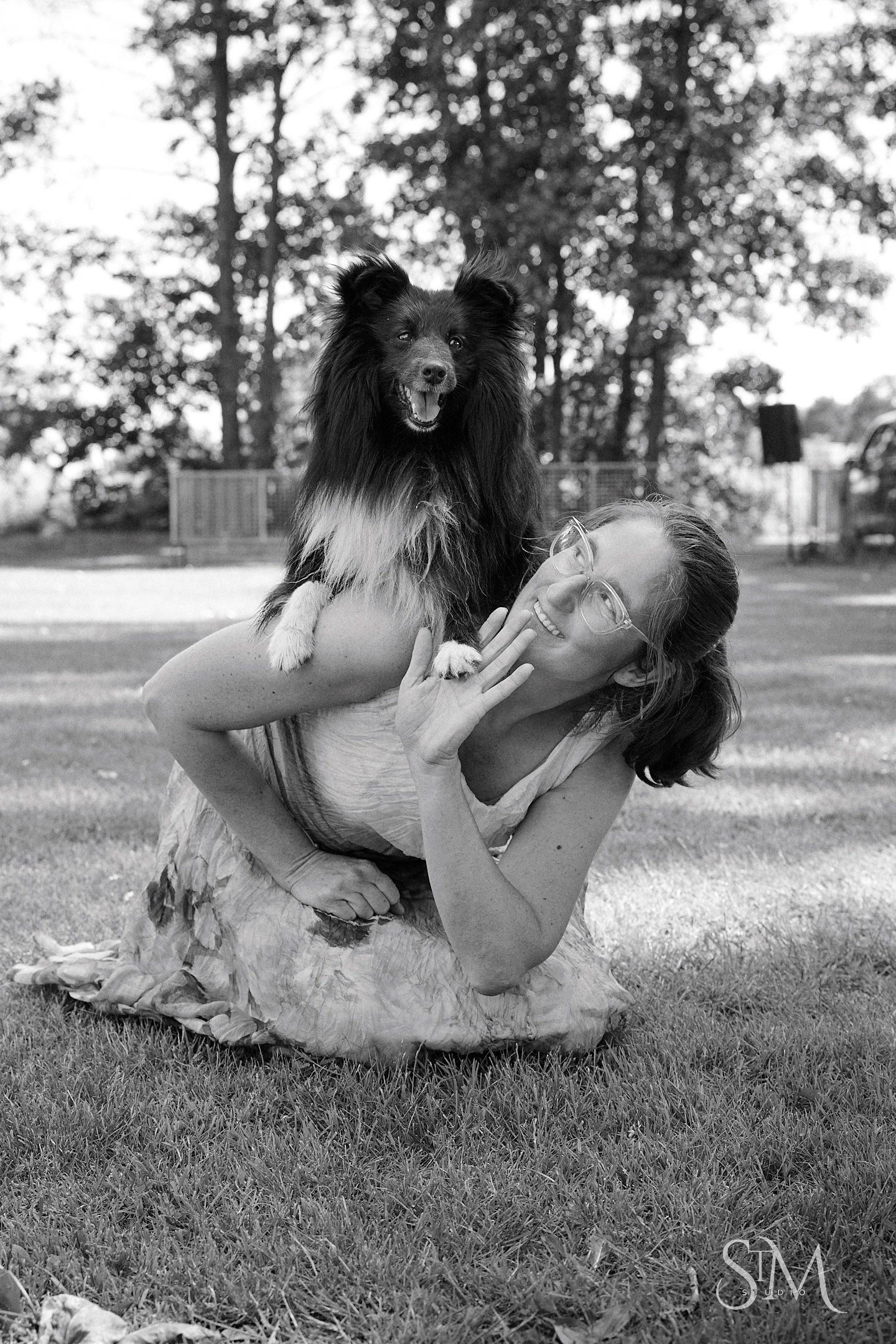 Black and white photo of a woman crouching on grass, lovingly holding a Shetland Sheepdog in an outdoor park setting.
