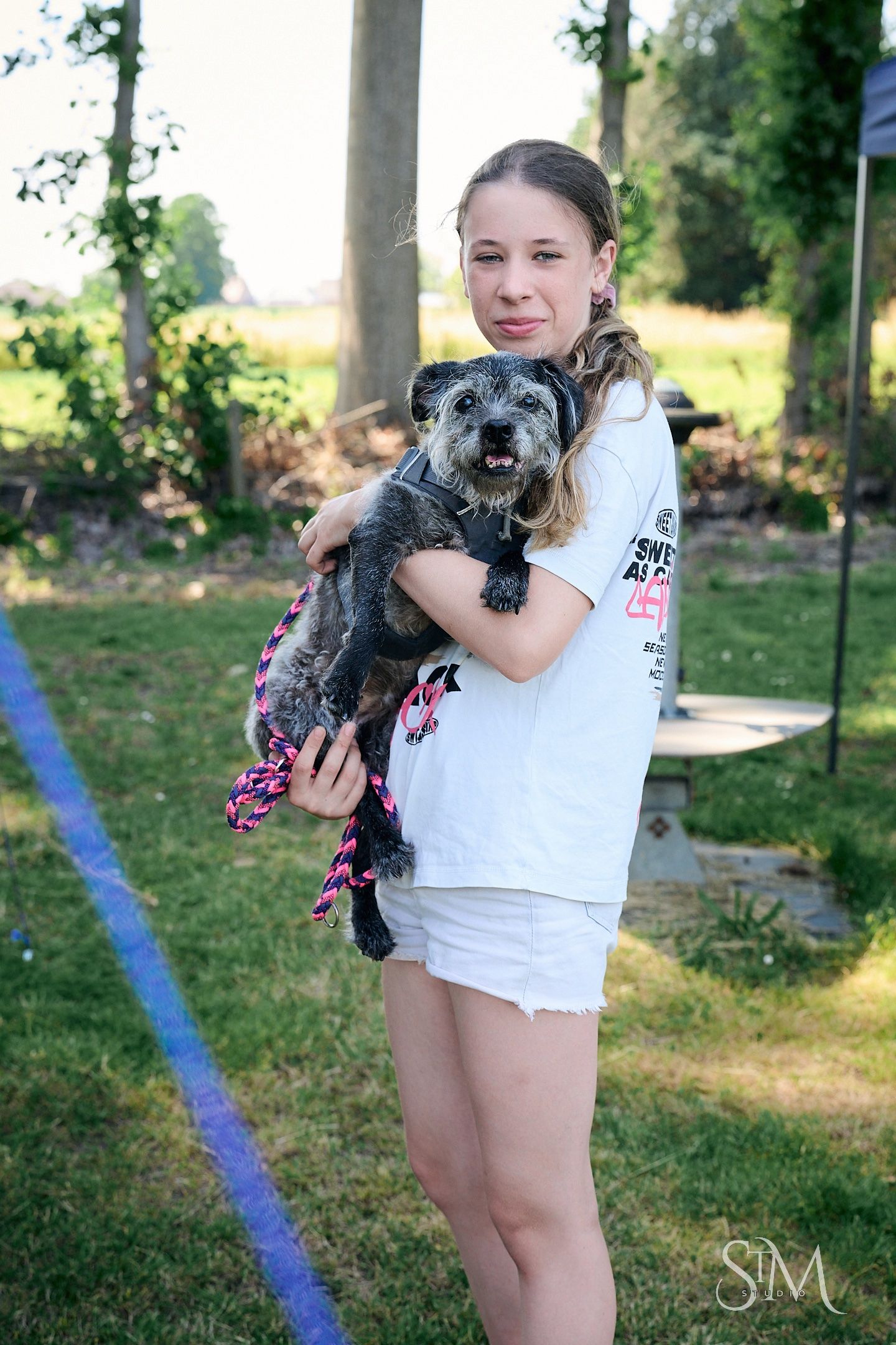 Young girl holding a small gray schnoodle dog outdoors on green grass, wearing white shirt and shorts.