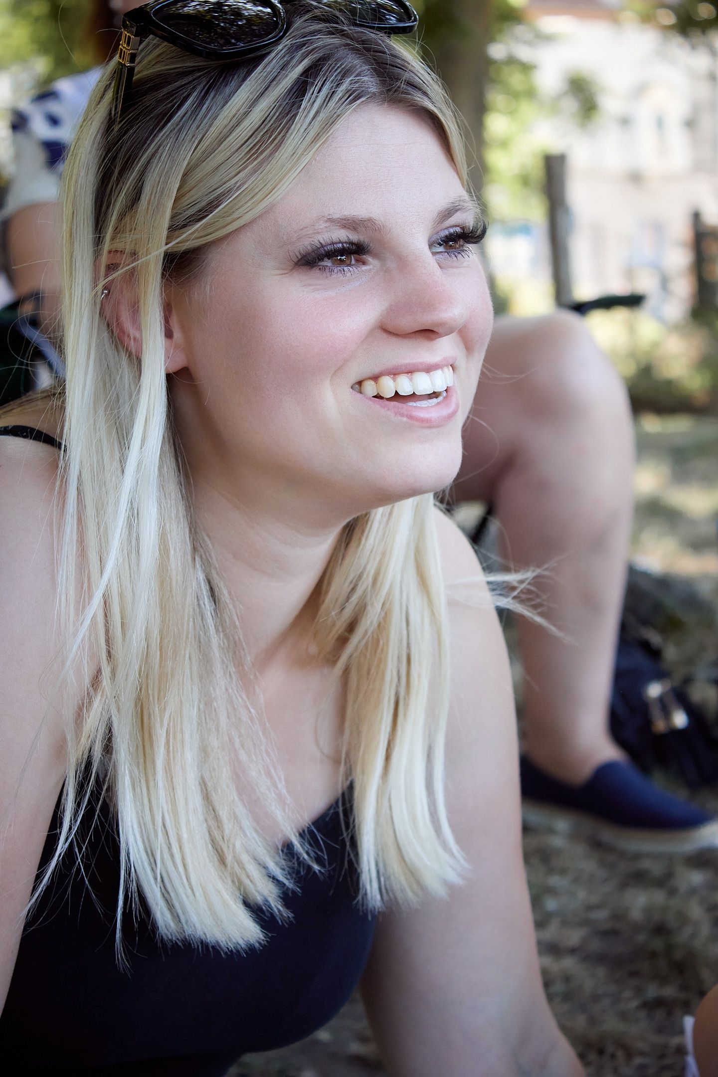 Smiling blonde woman with sunglasses on her head, wearing a black top, sitting outdoors on a sunny day.