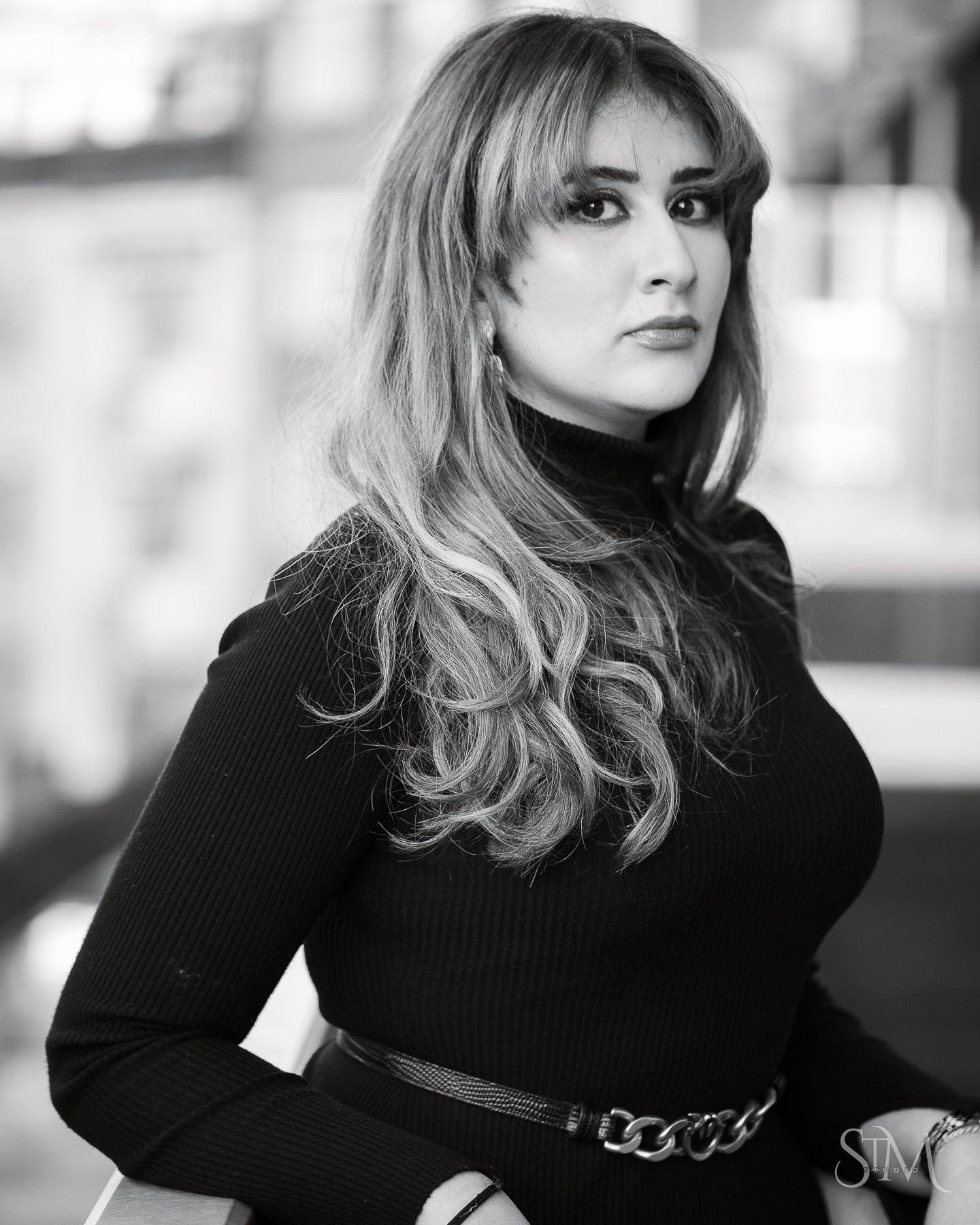 Black and white portrait of a woman with long wavy hair wearing a black turtleneck sweater and chain belt.