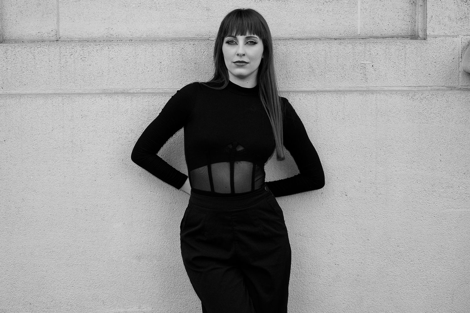 Black and white photo of a woman in a black sheer bodysuit and skirt posing against a light wall.