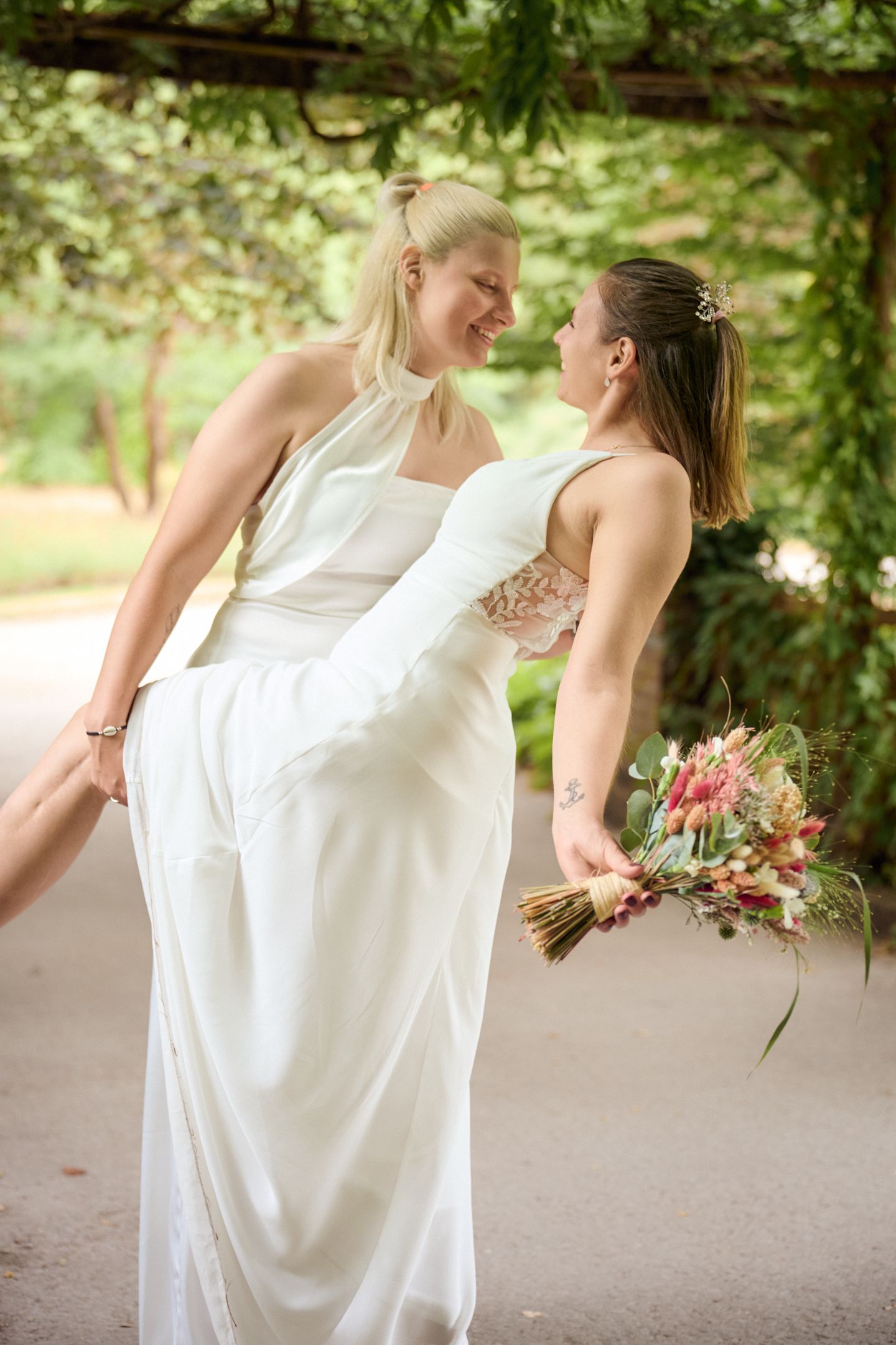 Two brides in white wedding gowns share an intimate moment, one holding a colorful bouquet under a leafy garden pergola.