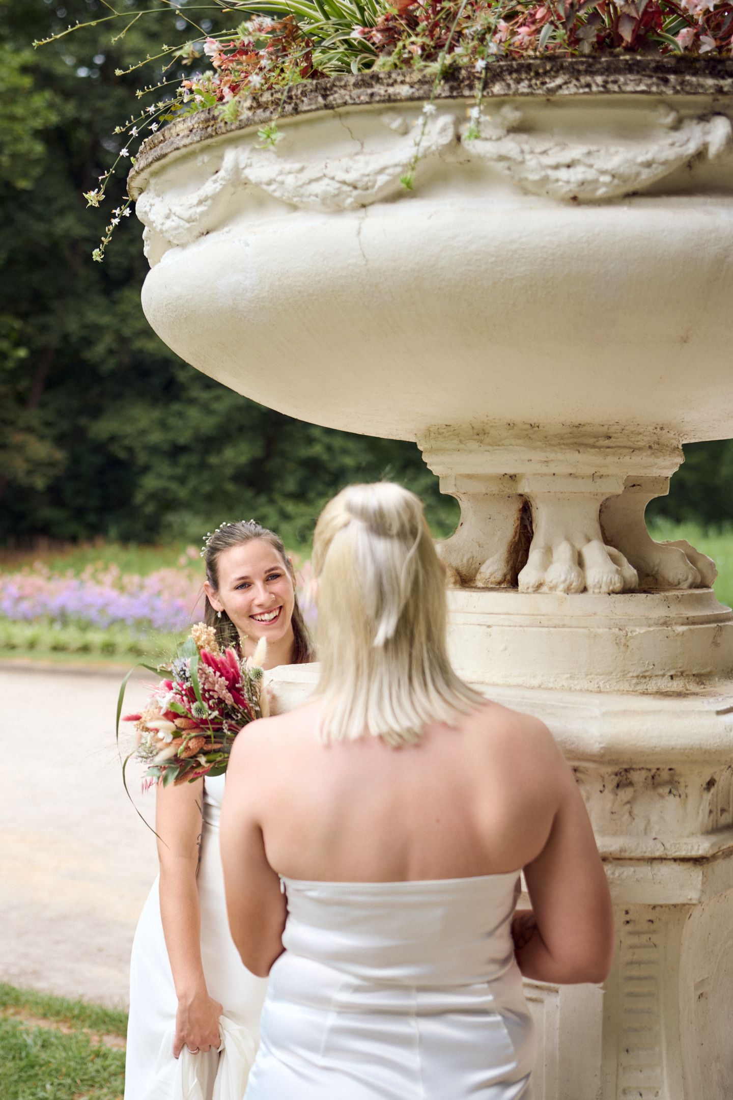 Two brides in white gowns share a joyful moment near an ornate stone urn planter in a lush garden setting.