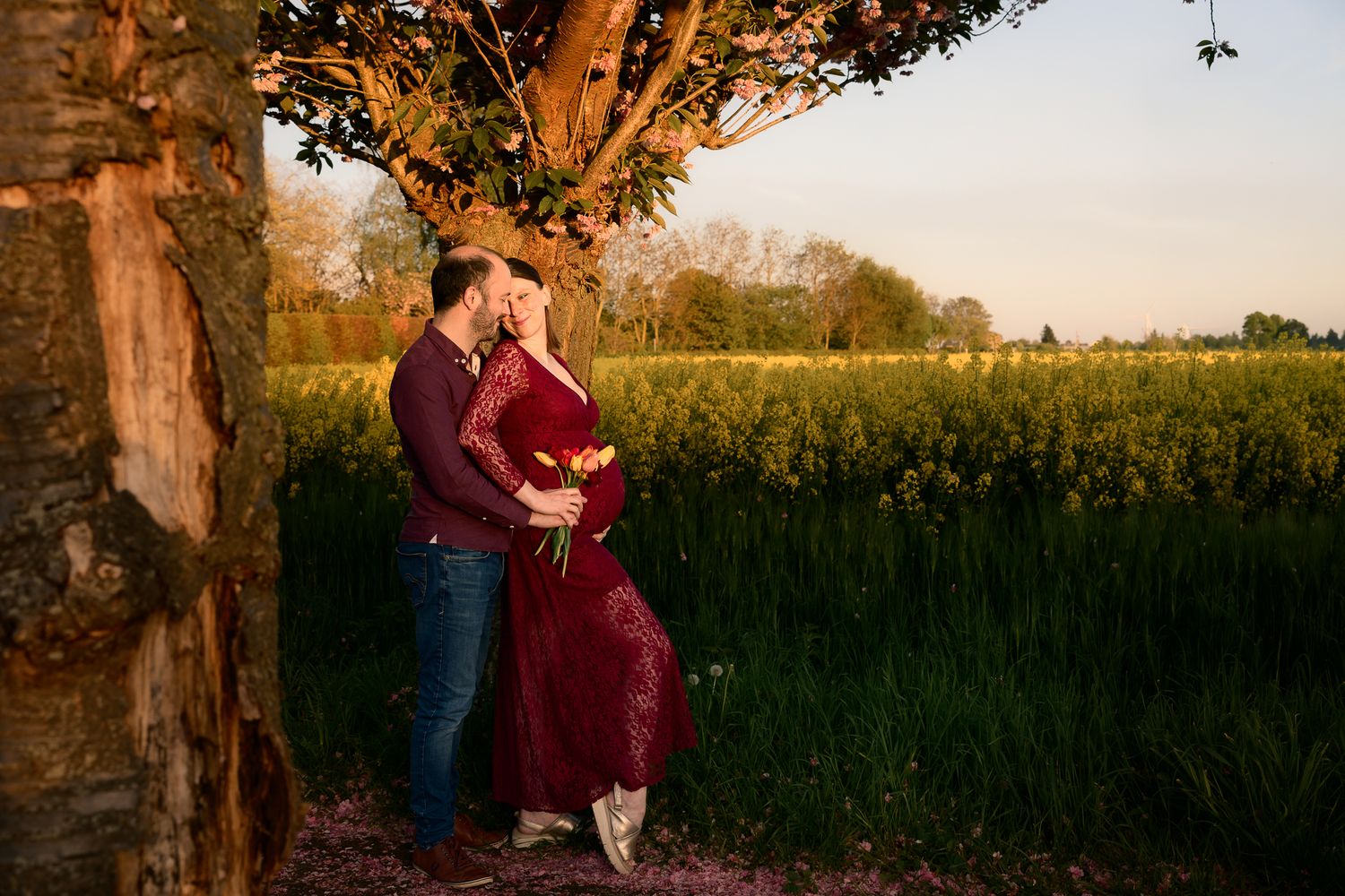 Couple embracing by a tree at golden hour near a vibrant green field, woman in red dress during maternity shoot.