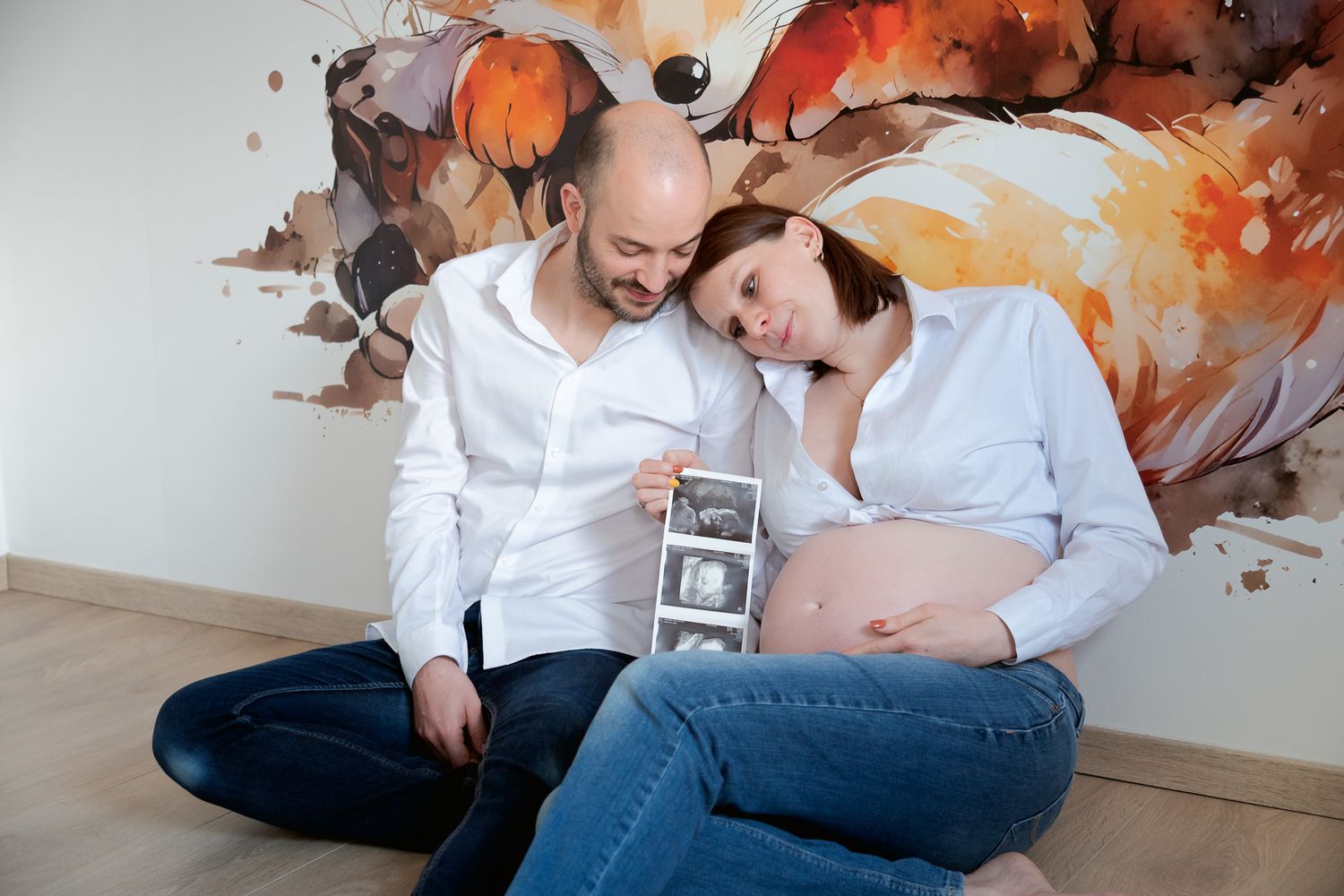 Expecting couple sitting on floor looking at ultrasound photos, pregnant woman in white shirt showing baby bump.