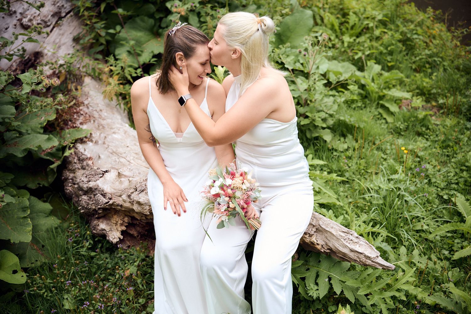 Two brides in white gowns share a romantic moment outdoors, surrounded by lush green foliage.