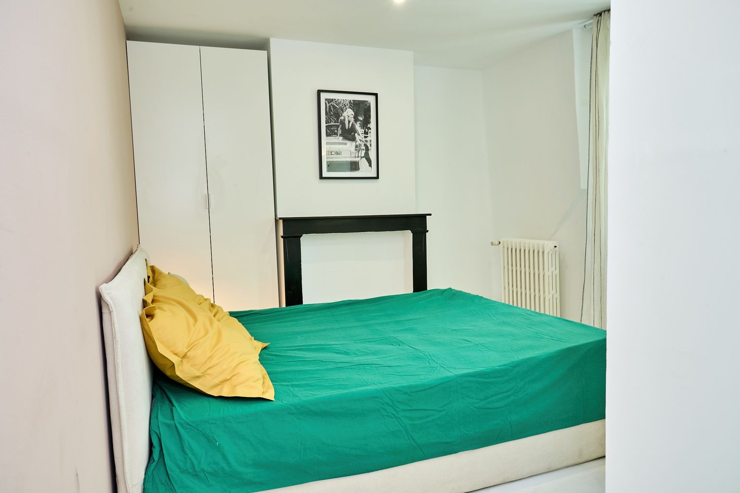 Cozy bedroom with teal bedspread, yellow pillows, white wardrobe, black fireplace, and framed artwork on white walls.