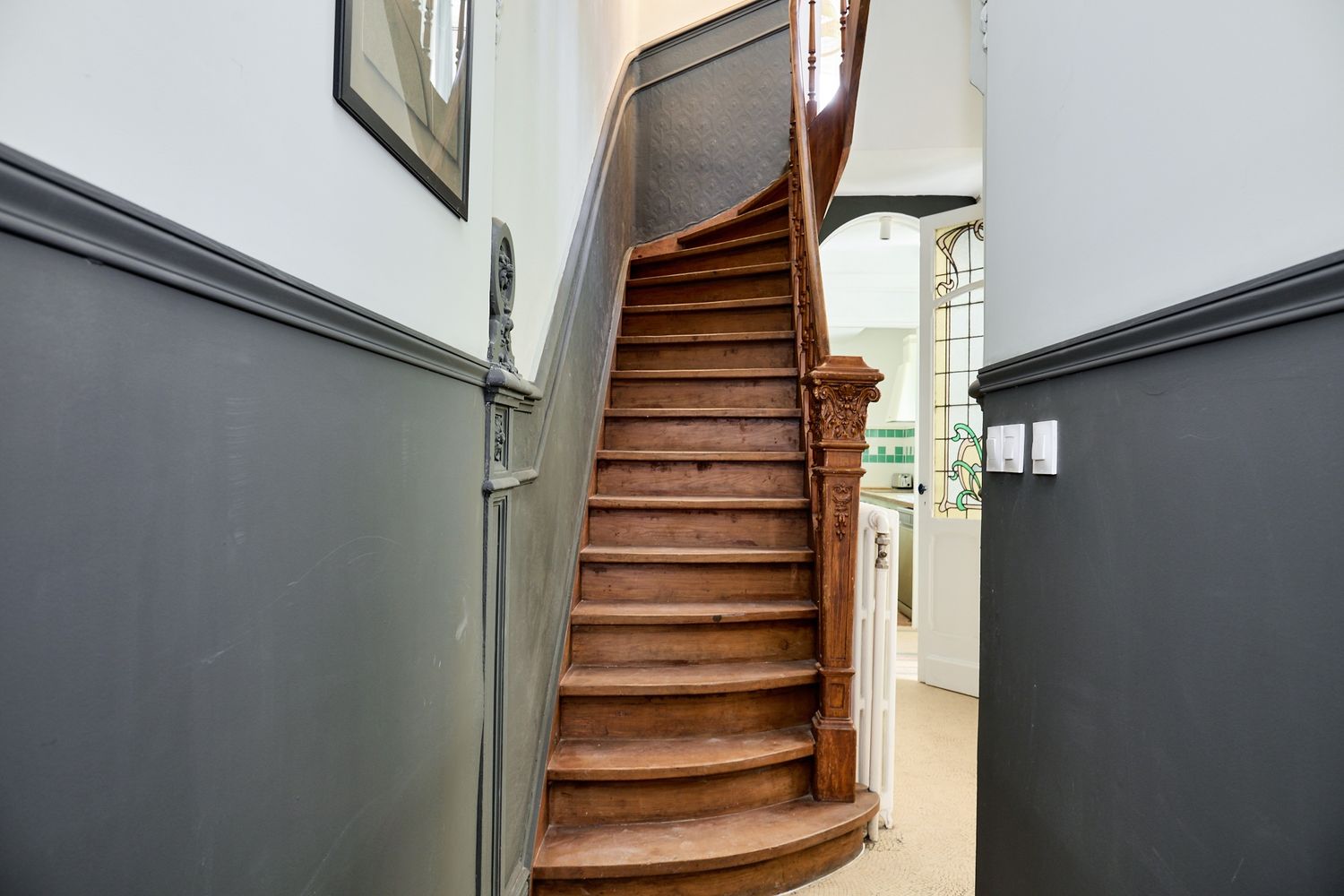 Elegant curved wooden staircase with ornate newel post in a classic hallway with grey wainscoting.