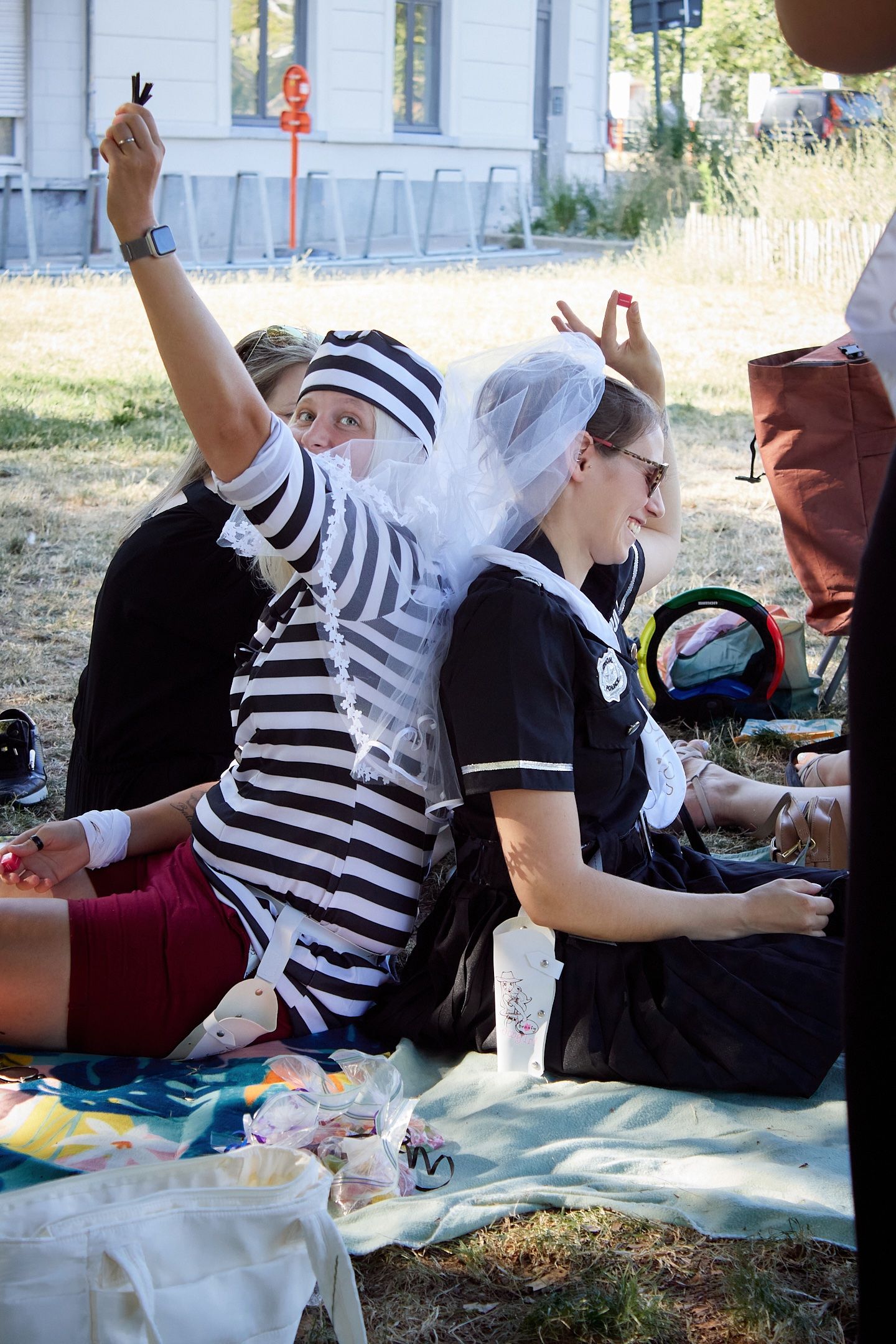 People in costumes sit outdoors on blankets, including a mime and bride, enjoying a festive gathering on a sunny day.