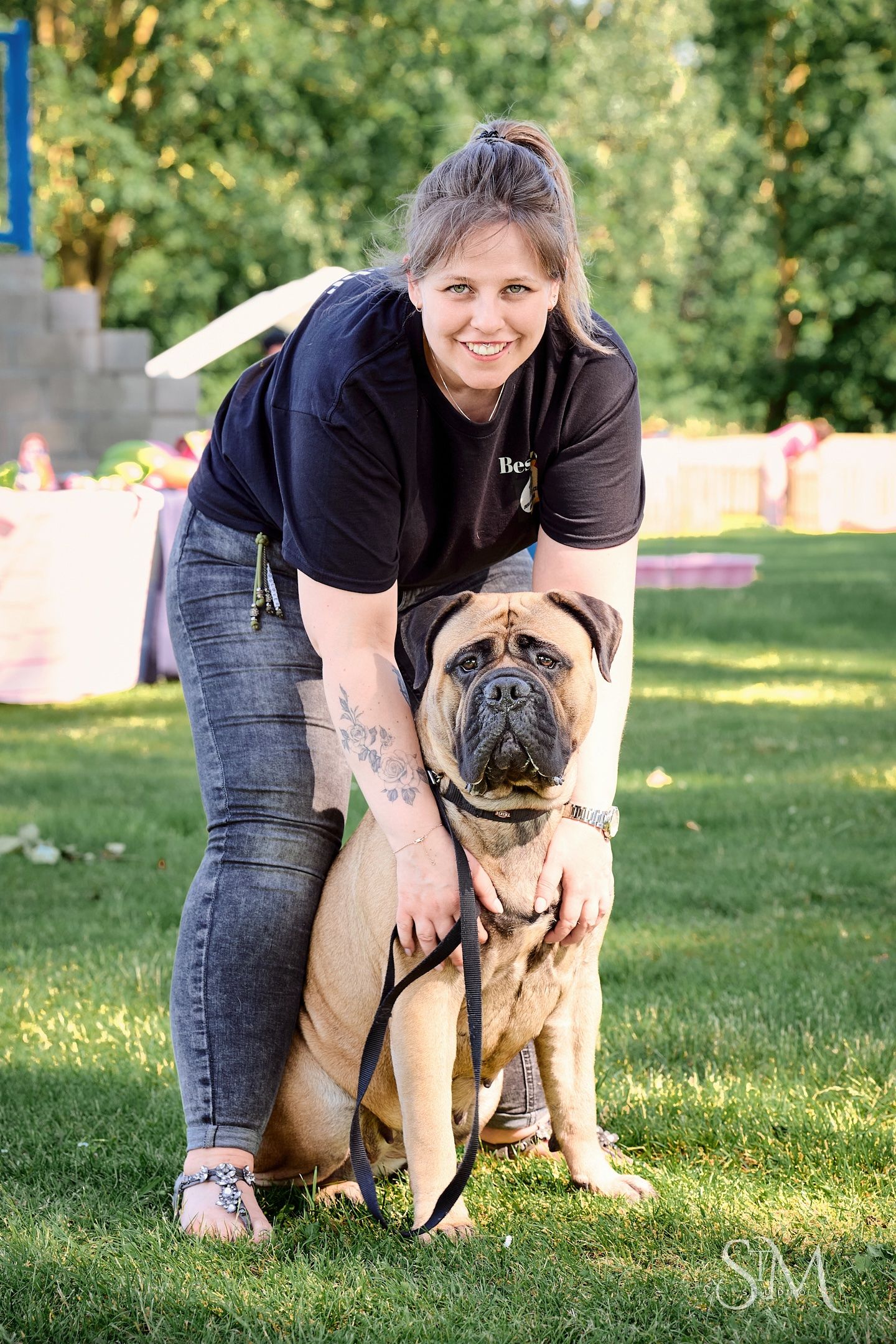 Woman in black shirt smiles while posing with her large Bullmastiff dog on a green lawn outdoors.
