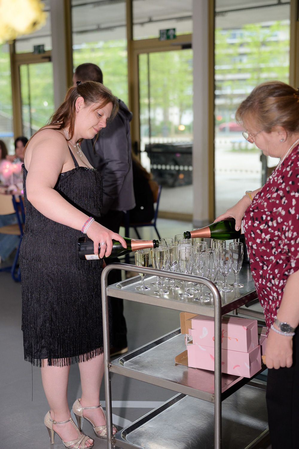 Woman in black fringe dress pouring champagne into glasses on a cart at a formal event.