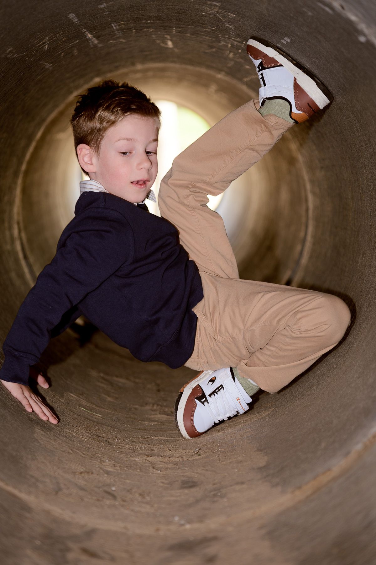 Young boy playing inside a large cylindrical tunnel, wearing khaki pants and white sneakers.