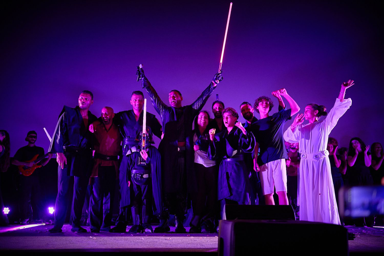 Group on stage with lightsabers under purple lights at a Star Wars themed live performance event.
