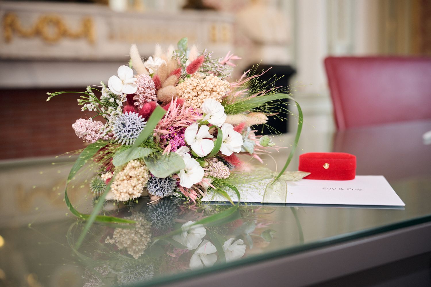 Elegant wedding bouquet with wildflowers beside a red ring box and card on a glass table.