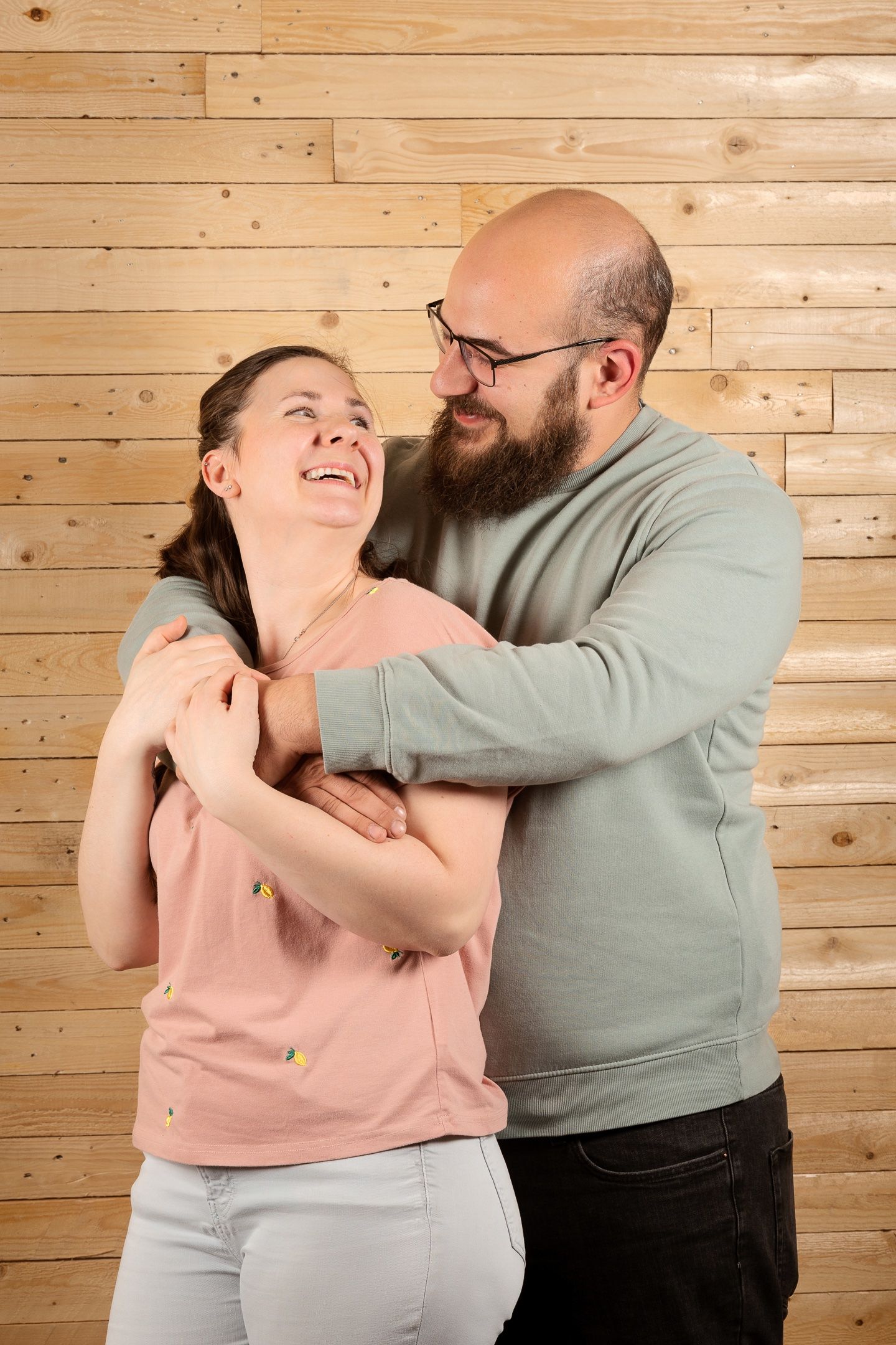 A bearded man in glasses hugs a laughing woman in a pink top against a wooden wall background.