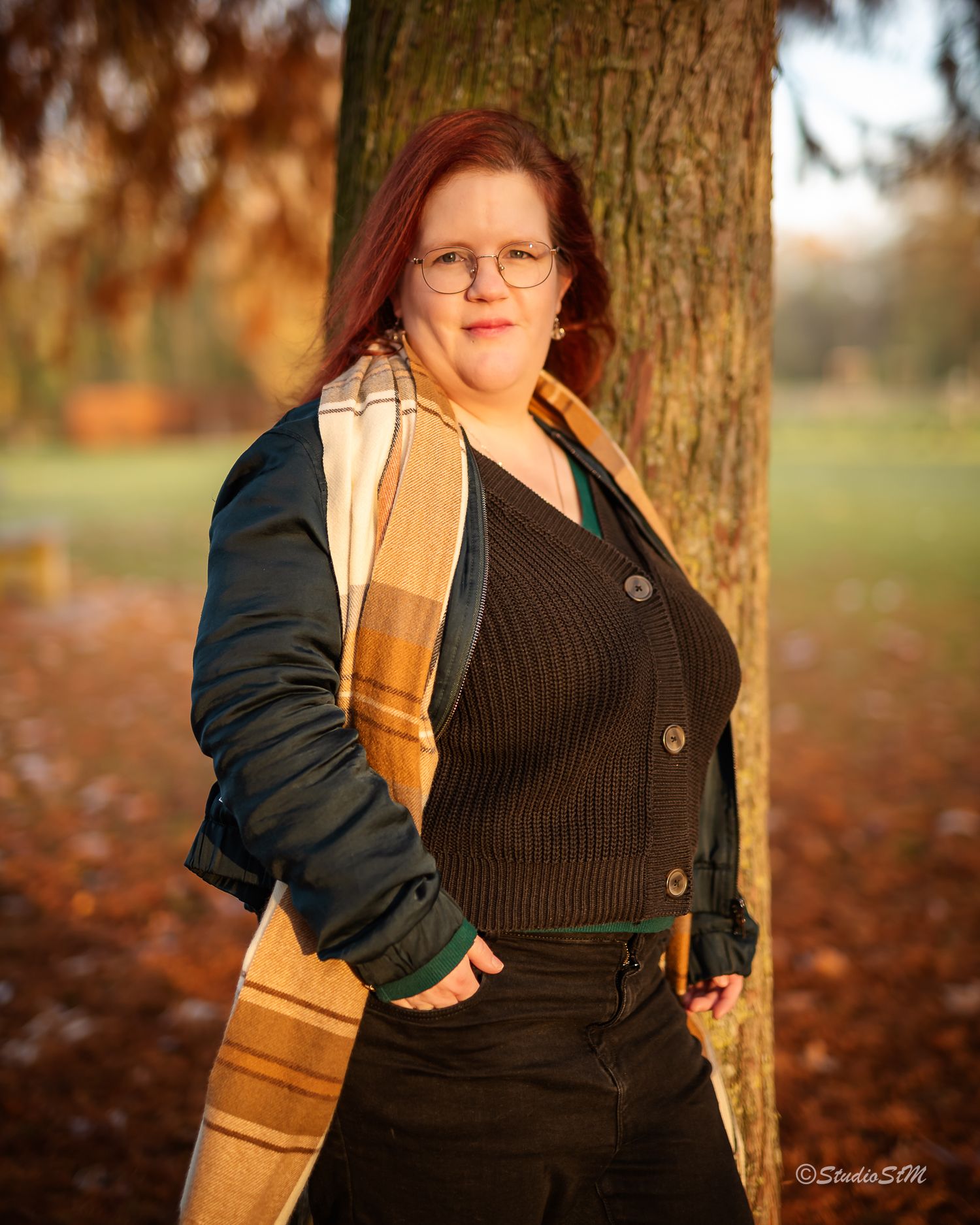 Red-haired woman in dark cardigan and plaid scarf leaning against tree in warm autumn sunlight.