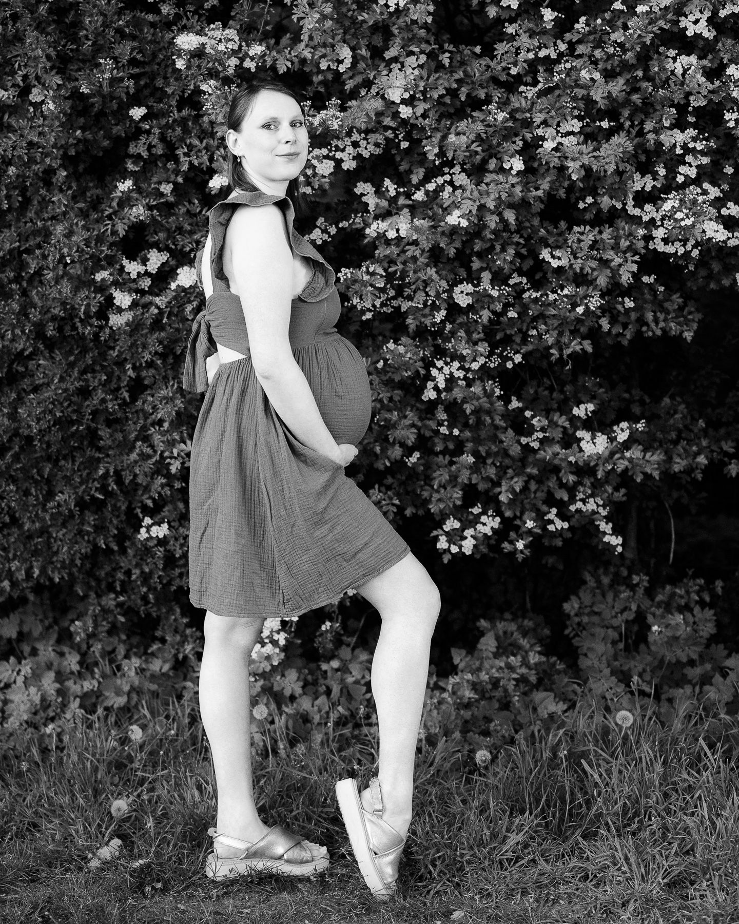 Pregnant woman in black dress and ballet shoes poses gracefully outdoors near blooming trees in black and white photo.